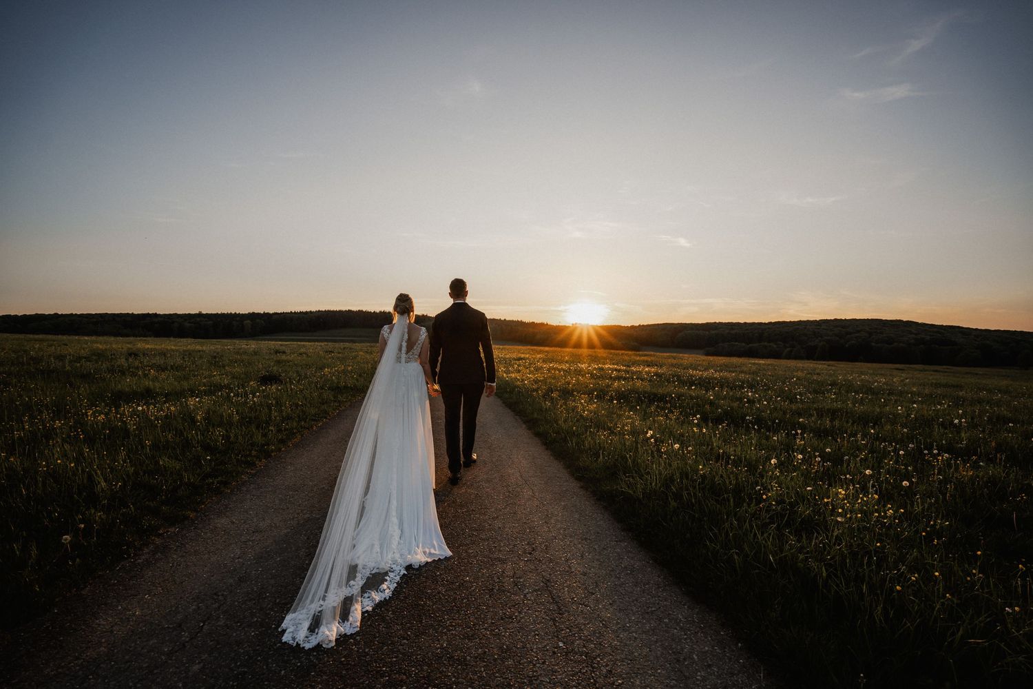 Two figures walk down a dirt road into the sunset with a flowing white wedding dress trailing behind.