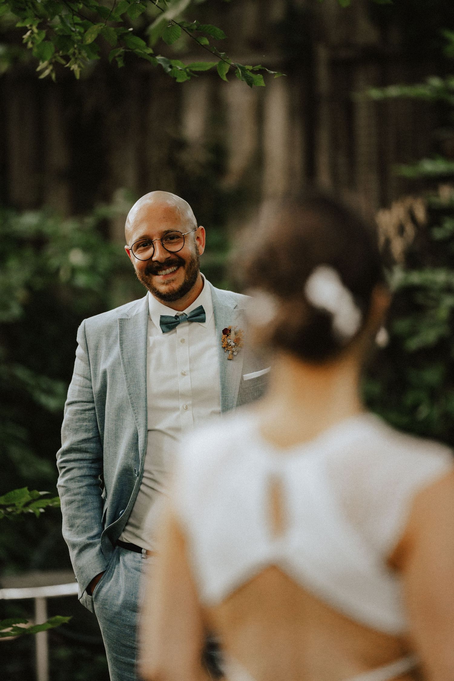 A groom in a light gray suit and bowtie smiles joyfully while looking at someone in a white wedding dress in a forest setting.