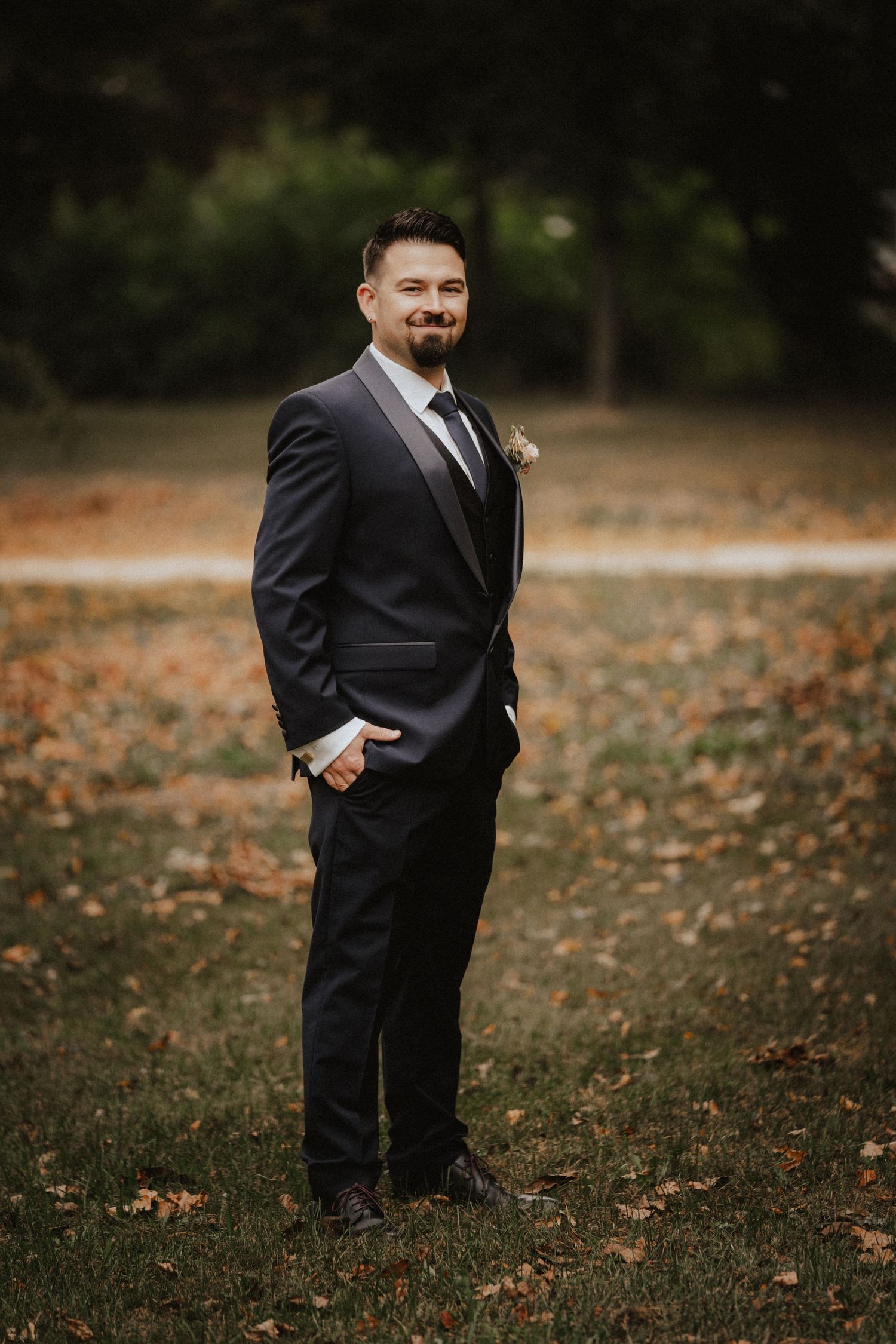 A figure in a dark formal suit stands in an autumn field covered with fallen leaves on a moody day.