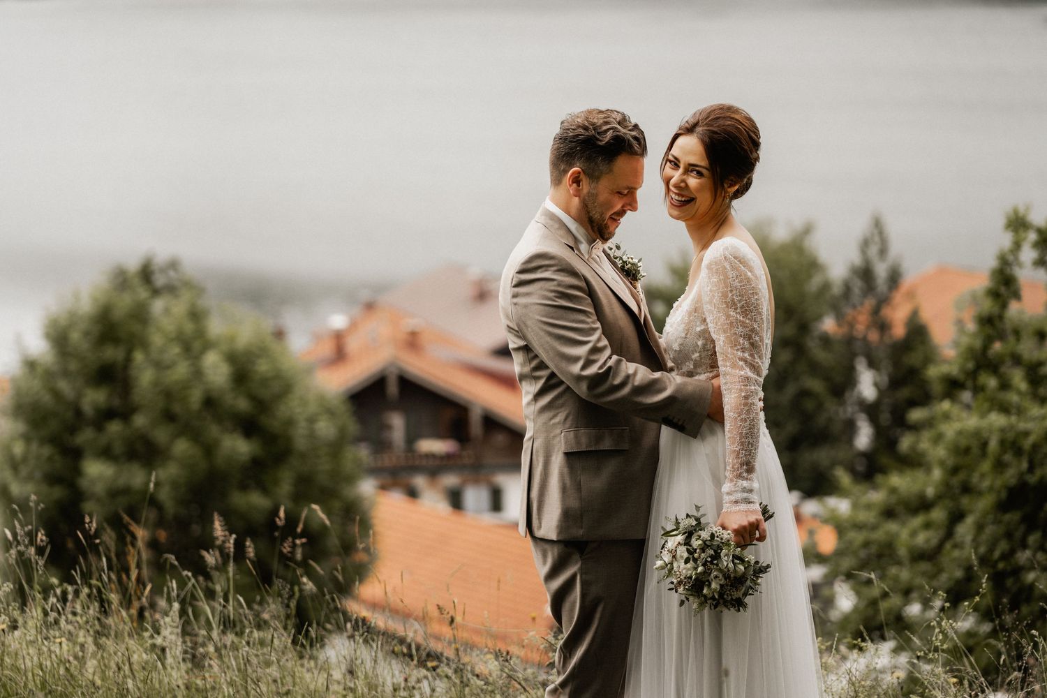 A bride and groom embrace outdoors with rustic mountain lodges and greenery in the background on their wedding day.