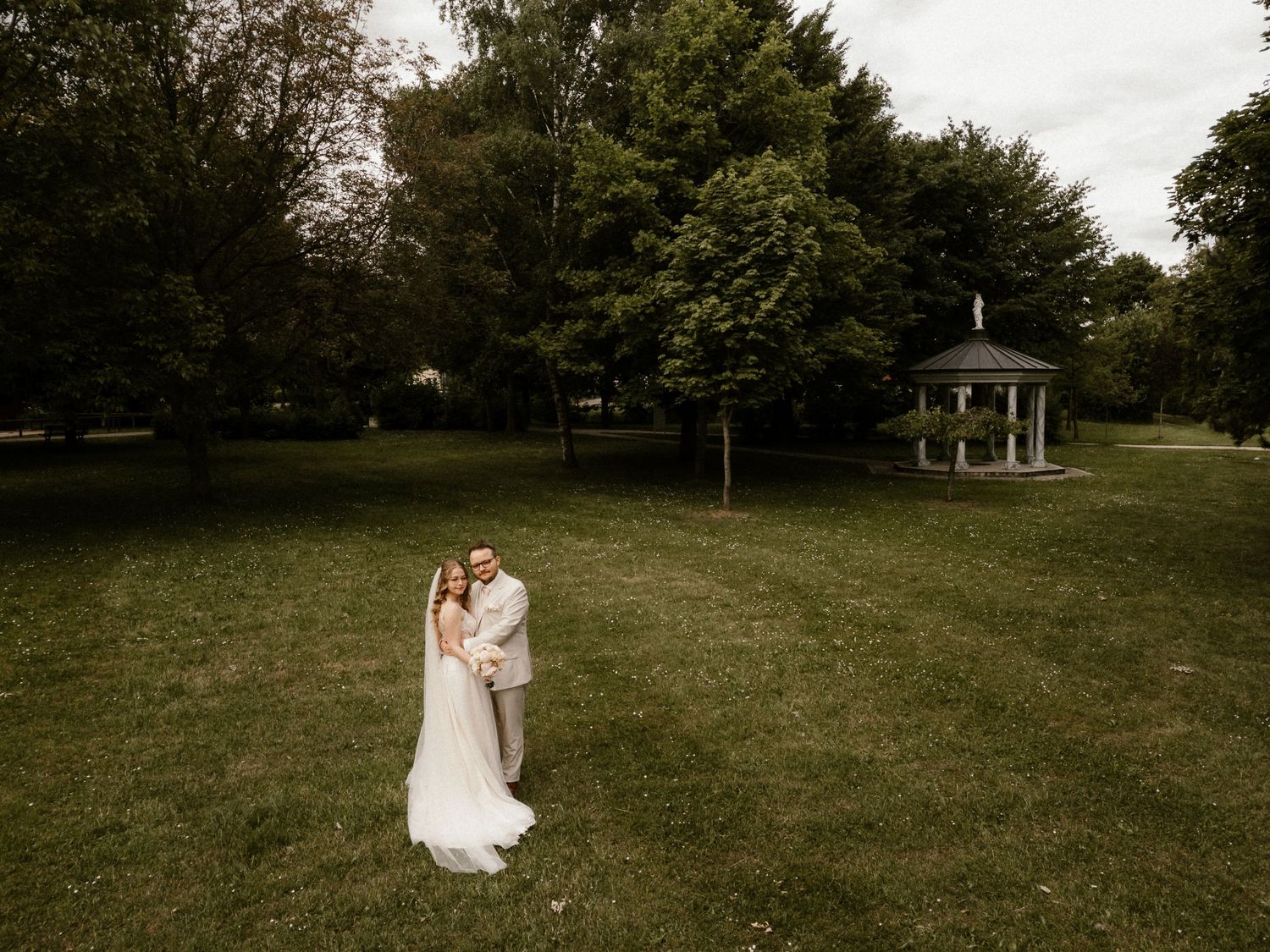 A couple stands together on a lush green lawn with a white gazebo visible in the background.