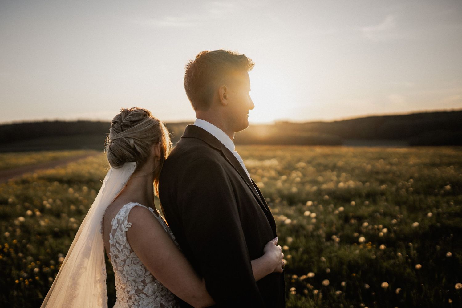 Bride and groom embrace in golden sunset light while standing in wildflower meadow during romantic outdoor wedding.