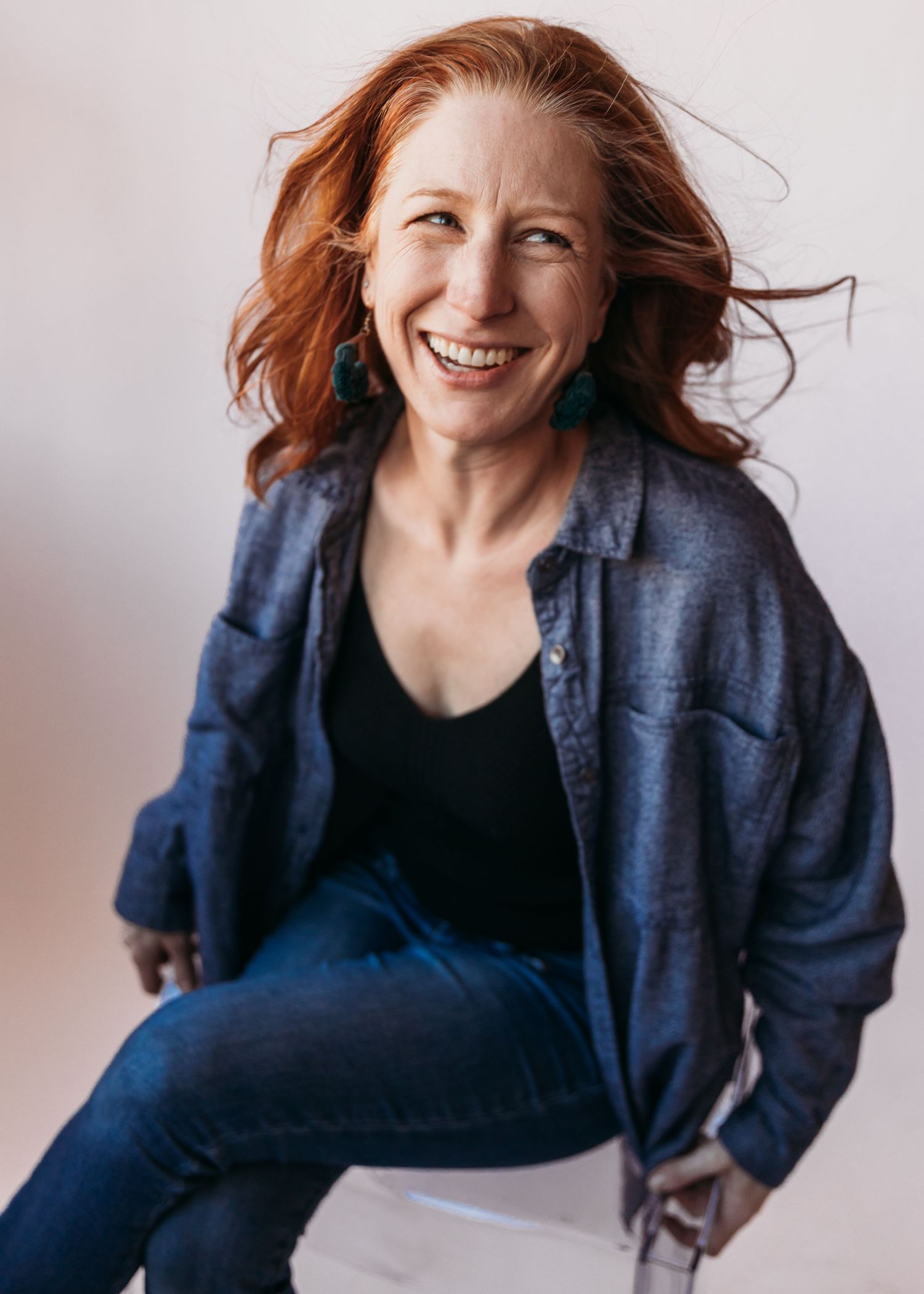 Happy woman with red windswept hair wearing a denim jacket and black top poses against white background.