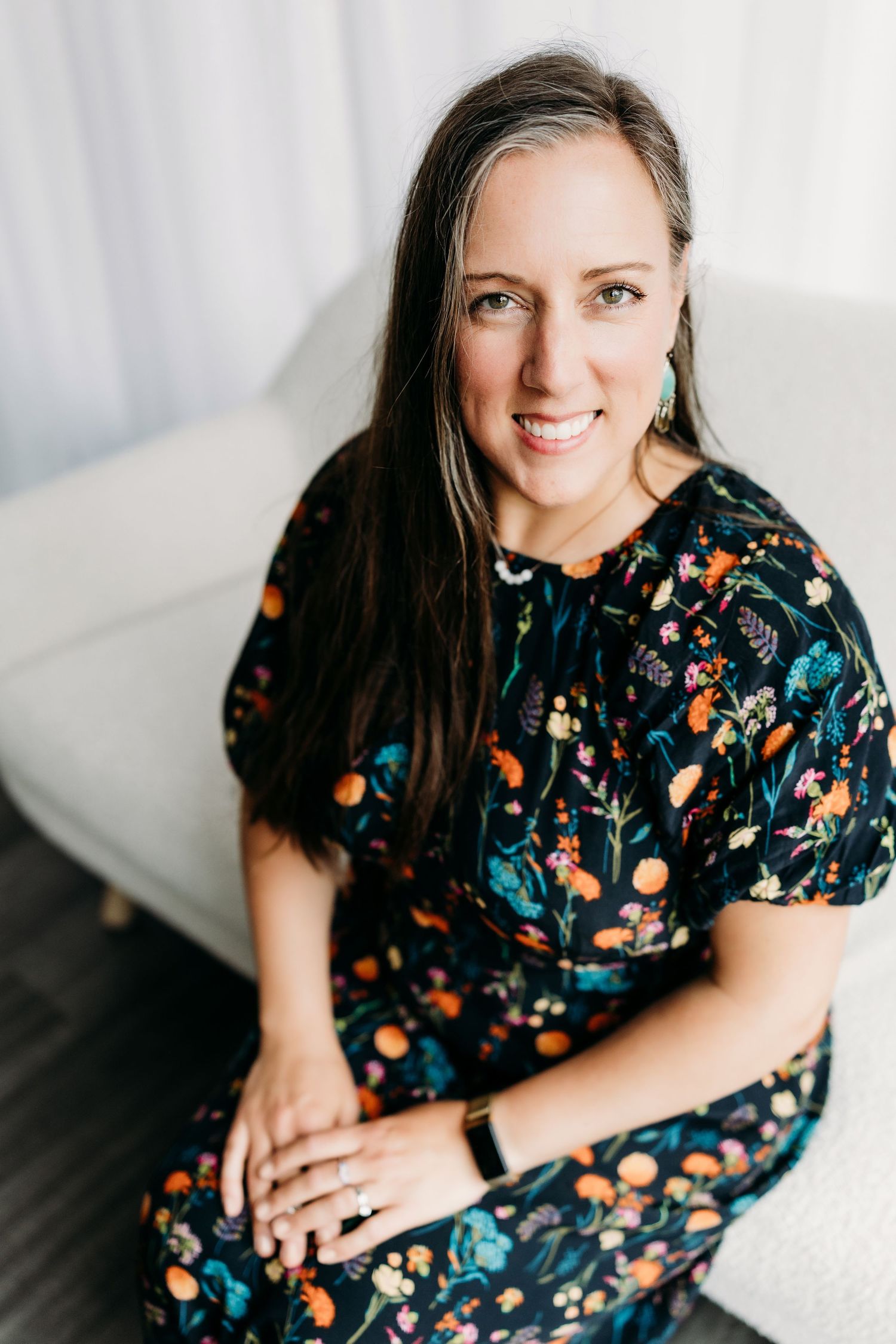 Portrait of someone wearing a dark floral dress with orange accents sitting against a white background.