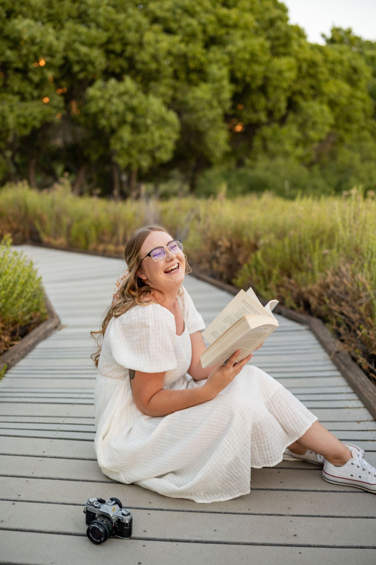 Senior in a white dress sits on a wooden boardwalk reading a book with a camera nearby on a sunny day in Jacksonville, NC.