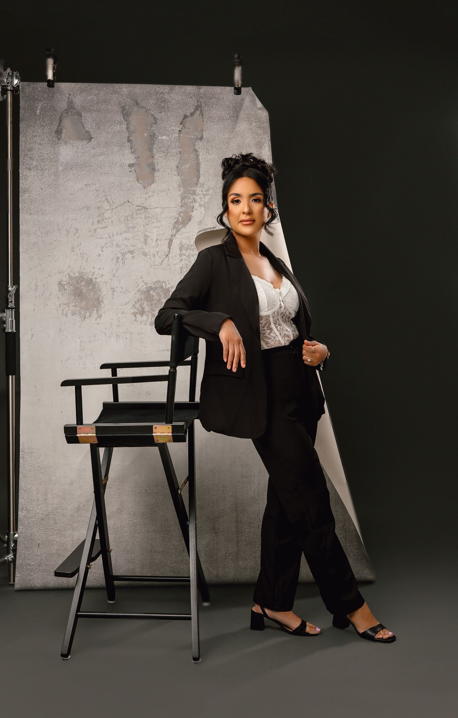 A person in a black blazer and pants leans elegantly against a director's chair in a studio setting with a textured backdrop.