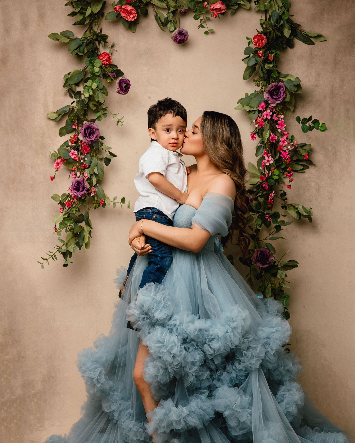 A dreamy portrait against a floral arch backdrop featuring a flowing dusty blue gown and tender embrace.