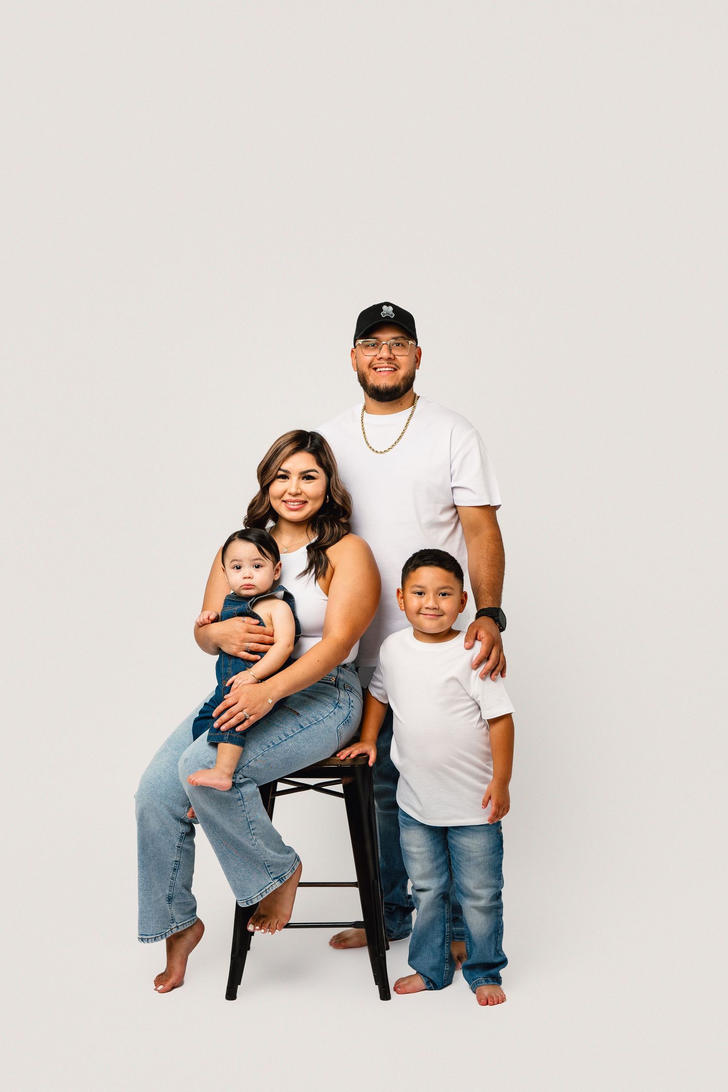 Family in white shirts and jeans poses together against white background, with one person seated on black stool.