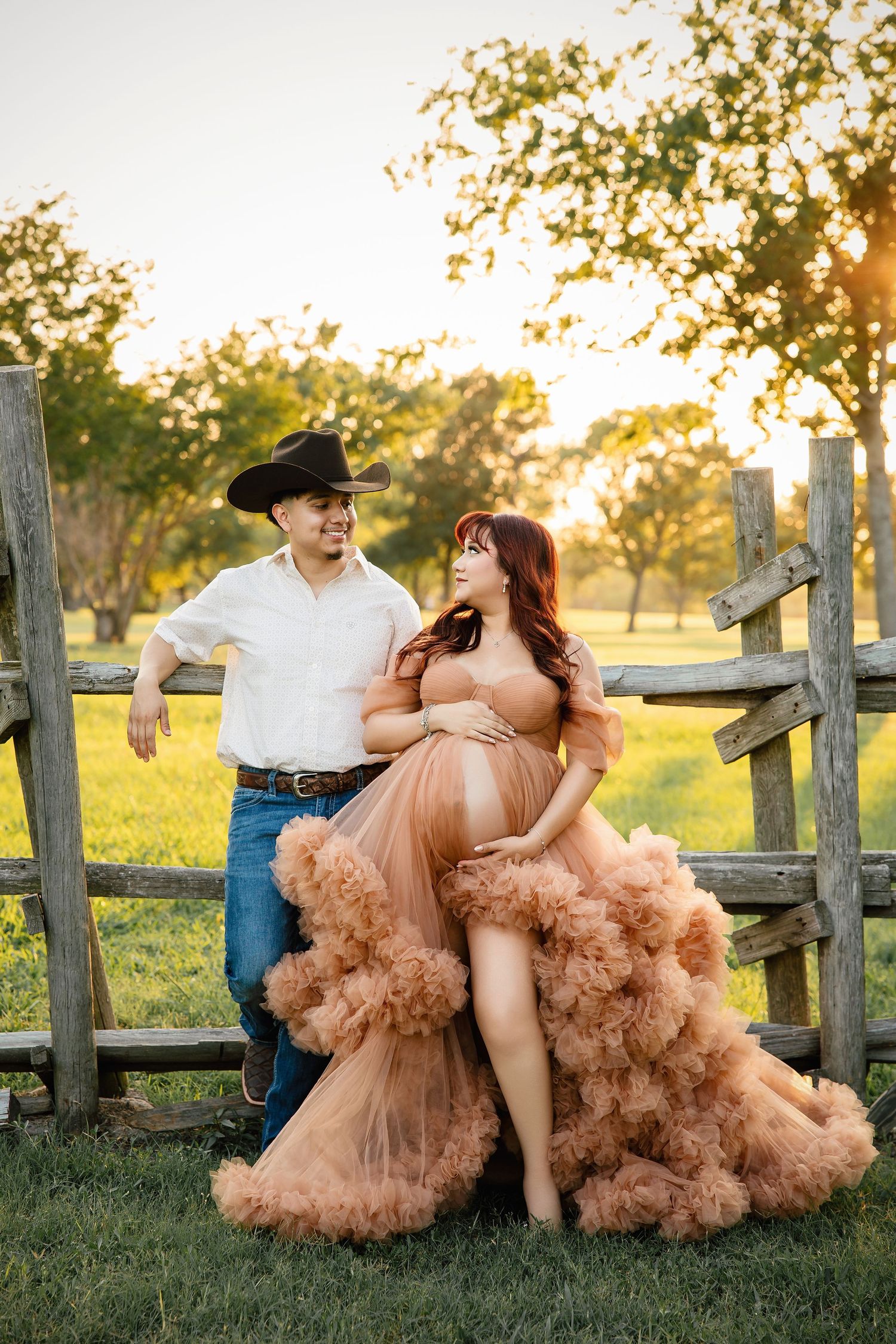 A couple sits playfully on a rustic wooden fence at sunset as her peachy tulle dress billows in the wind.