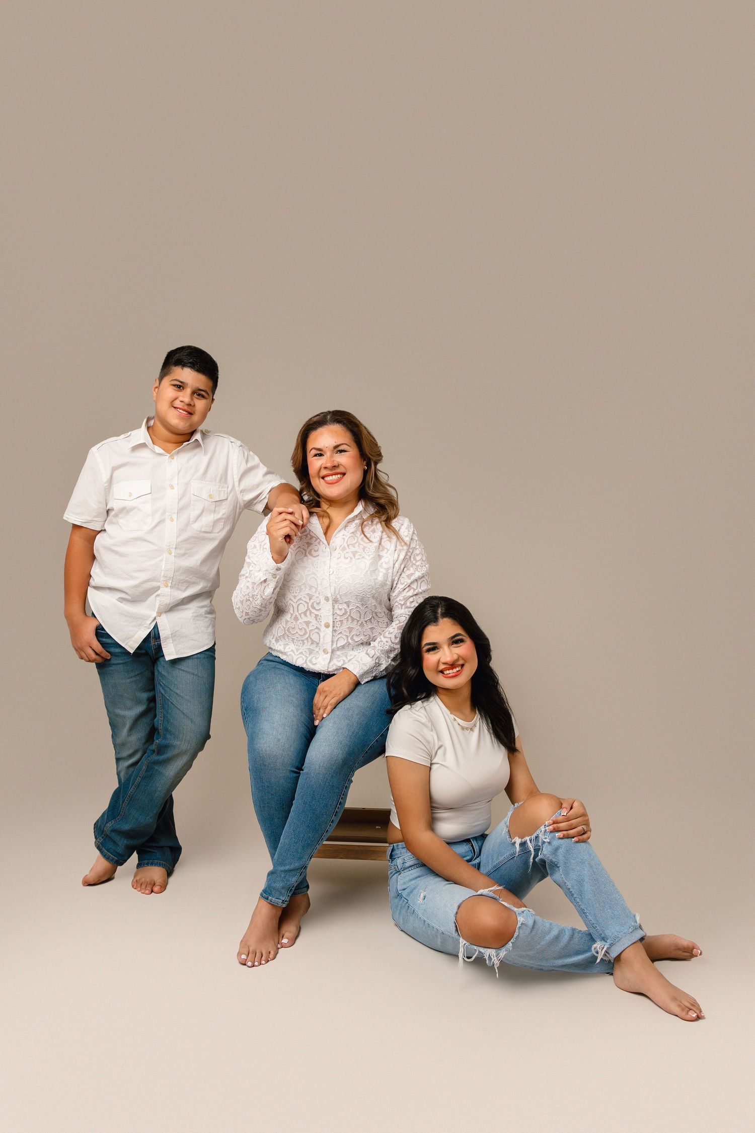 Three people in coordinated white tops and blue jeans pose casually against beige background in studio setting.