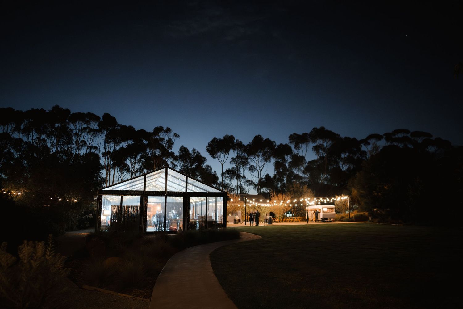 A white barn-style wedding venue illuminated at night with string lights and surrounded by tall trees.