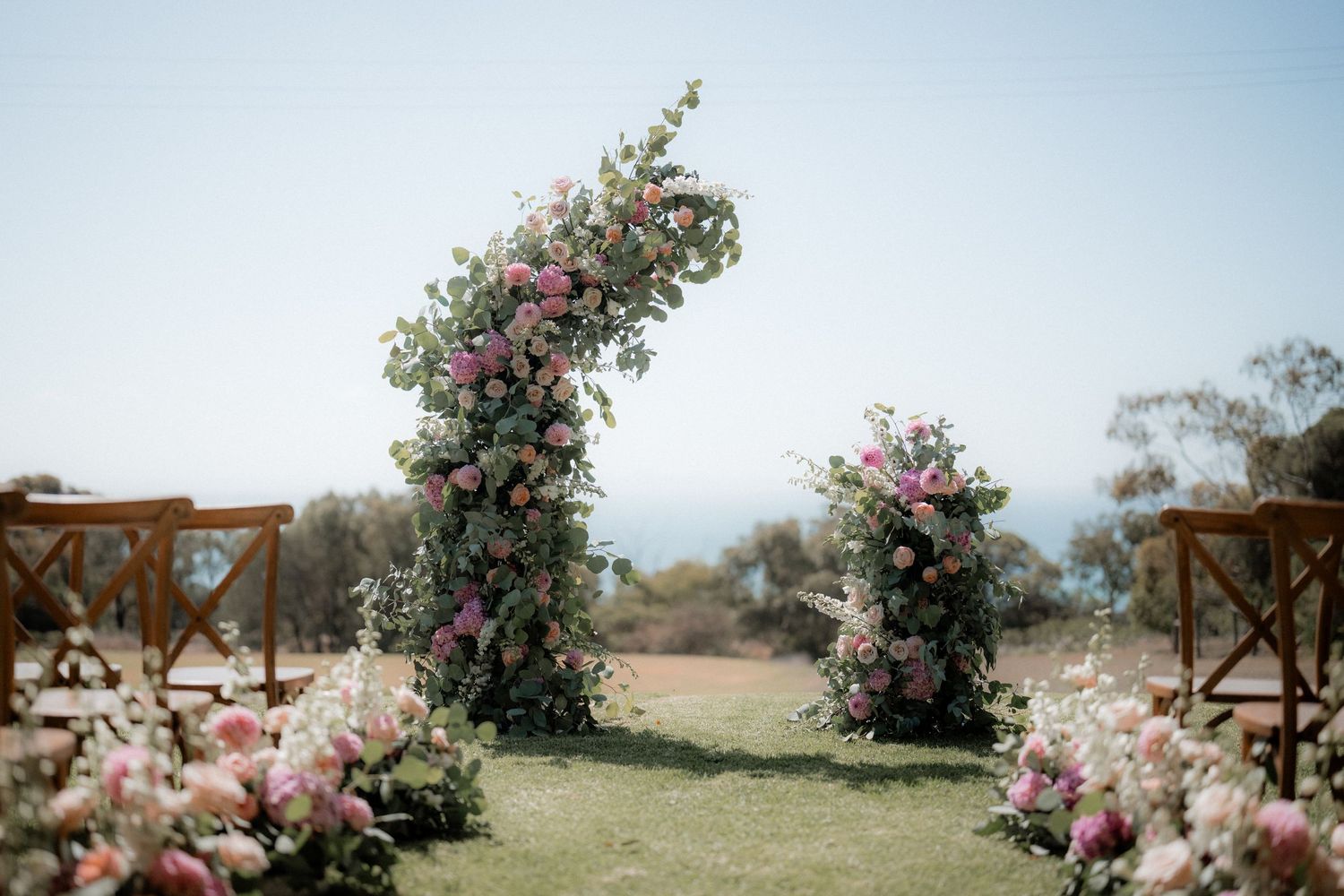 Two floral arches with pink and white blooms frame an outdoor wedding ceremony space.