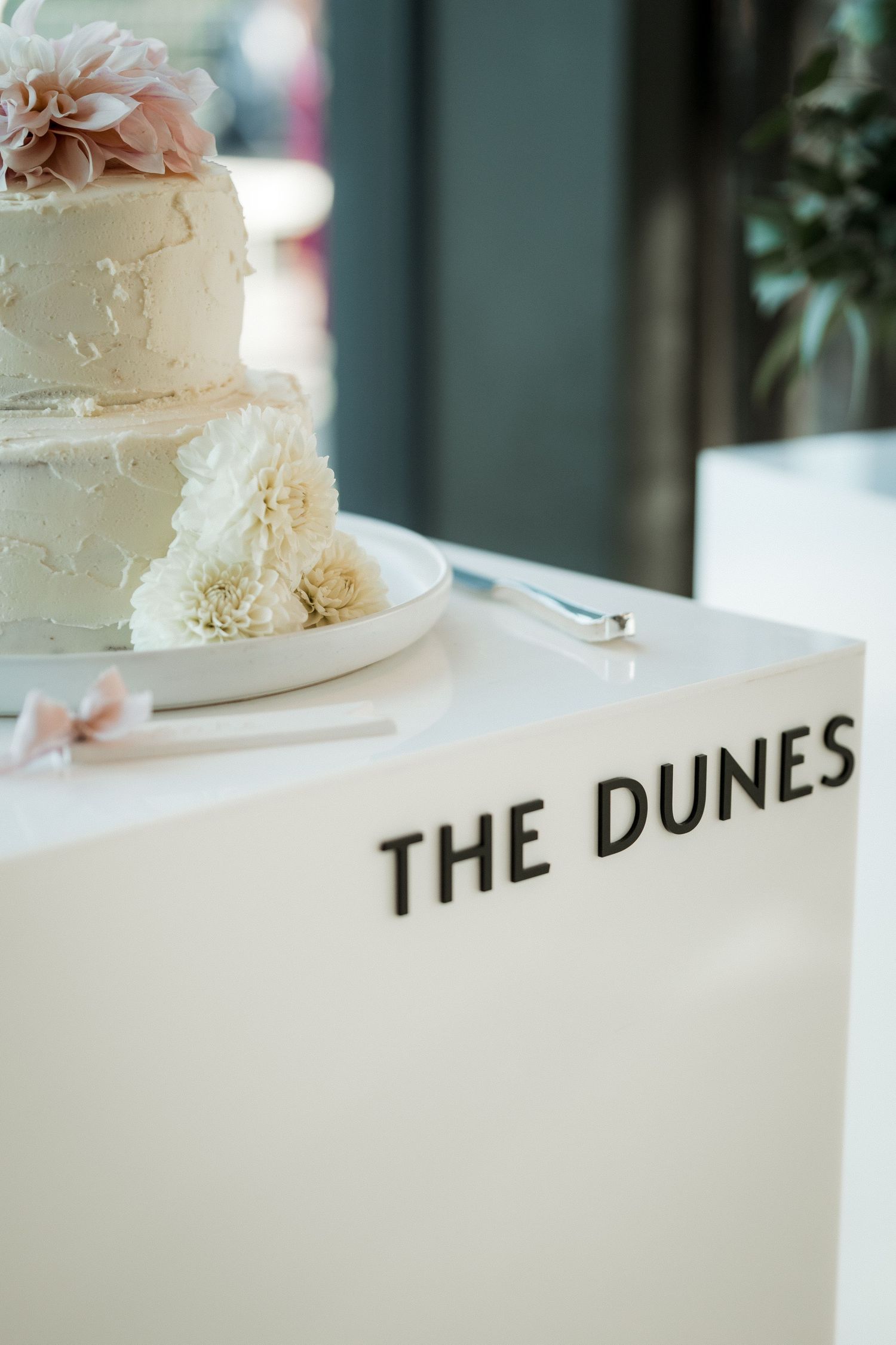 An elegant white wedding cake with floral decorations sits on a table labeled 'The Dunes'.