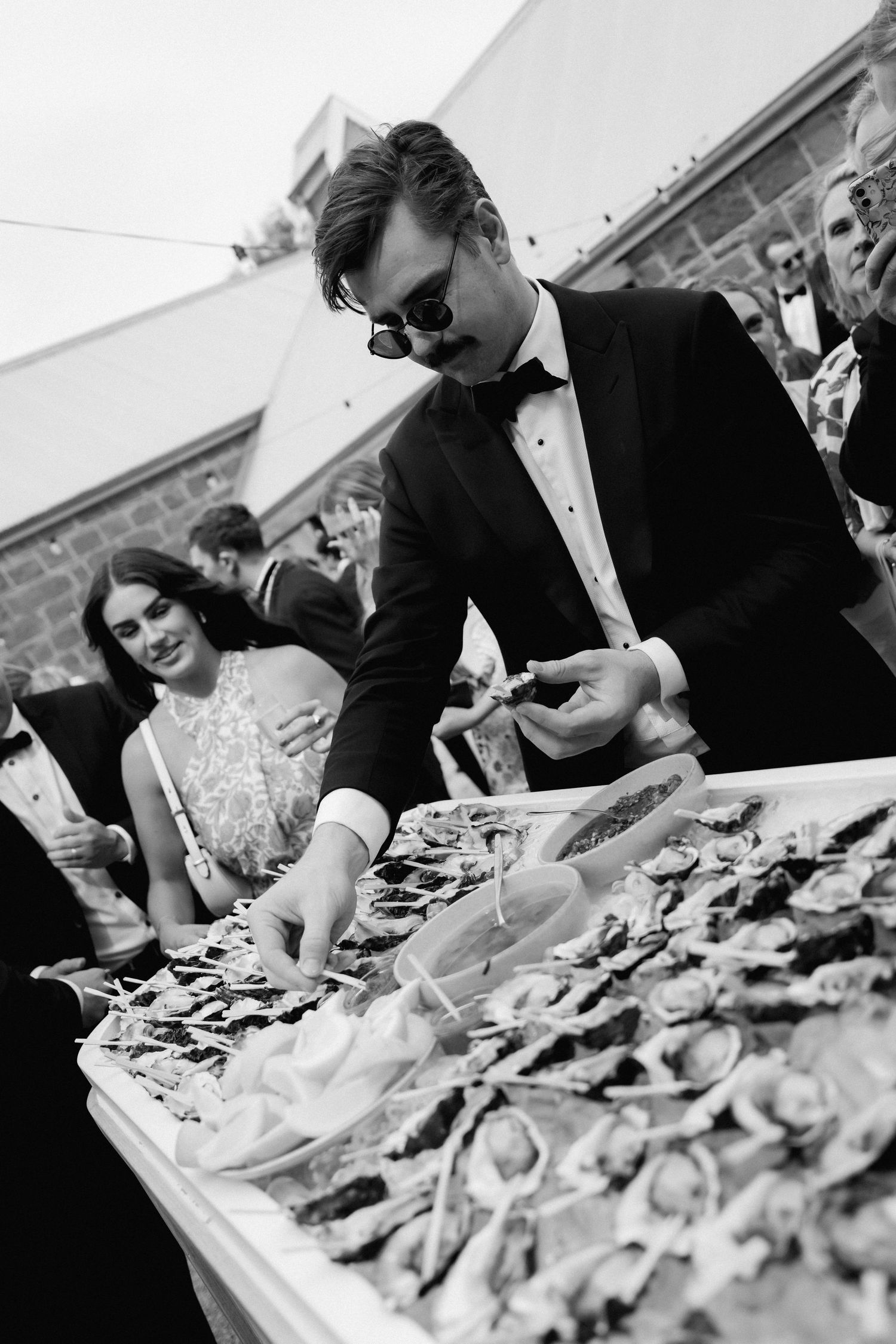 A formal event guest in sunglasses selecting oysters from a seafood display.