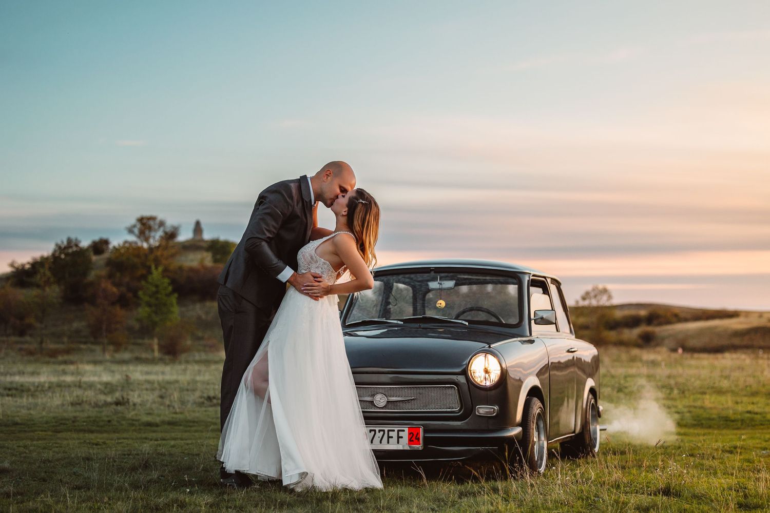 Klasszikus Trabant autó előtt egy esküvői pár áll, a naplemente fényeivel a háttérben, egy vidéki úton.