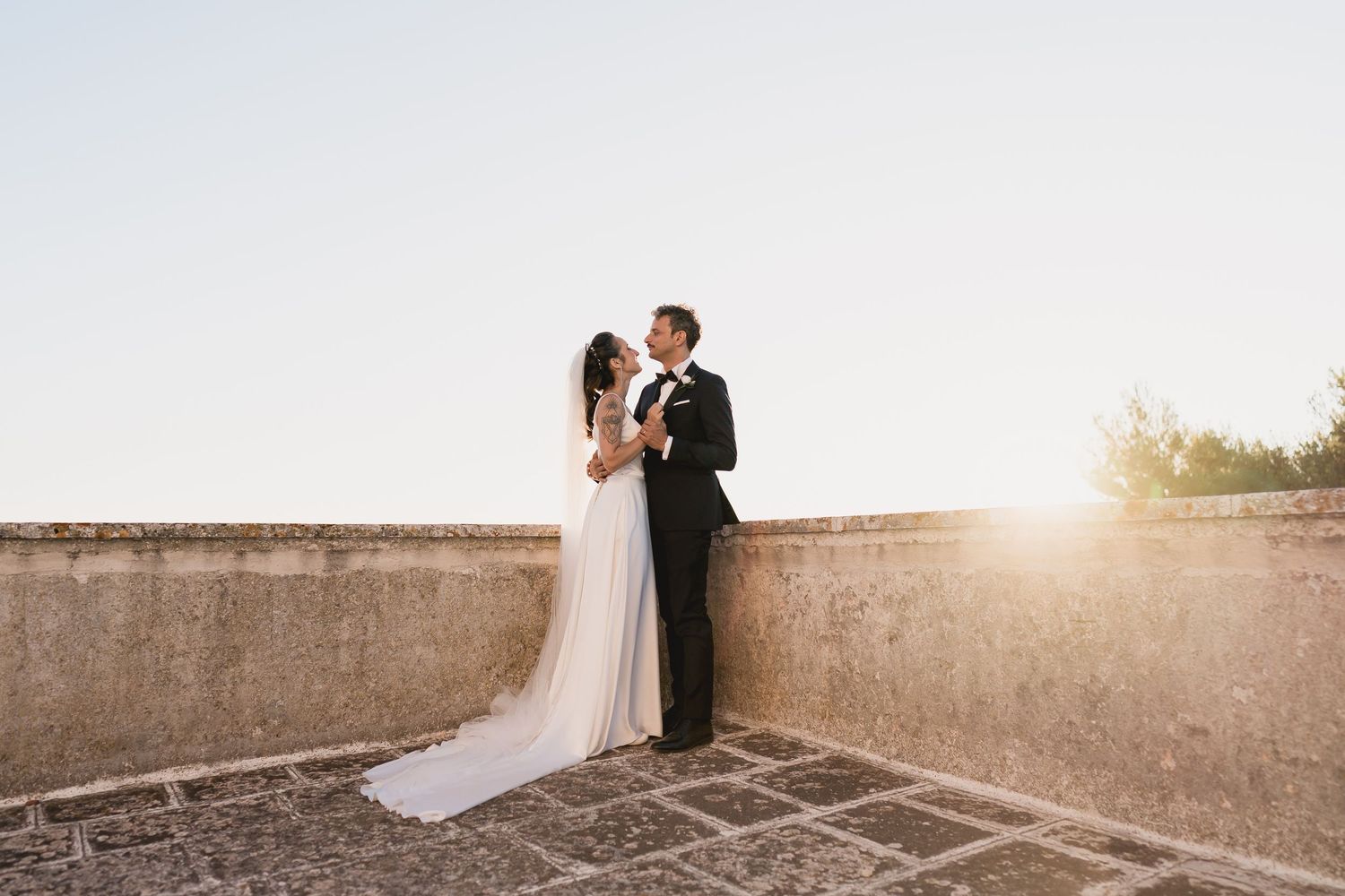 Two newlyweds embrace on a stone terrace during a romantic golden hour sunset.