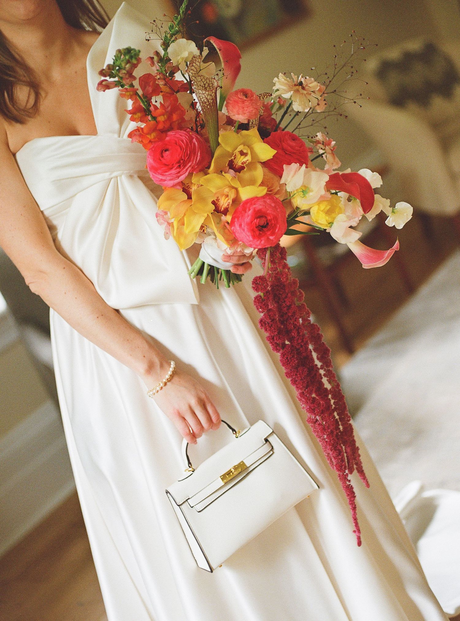 Person in white dress holding white designer handbag with vibrant coral and yellow floral bridal bouquet.