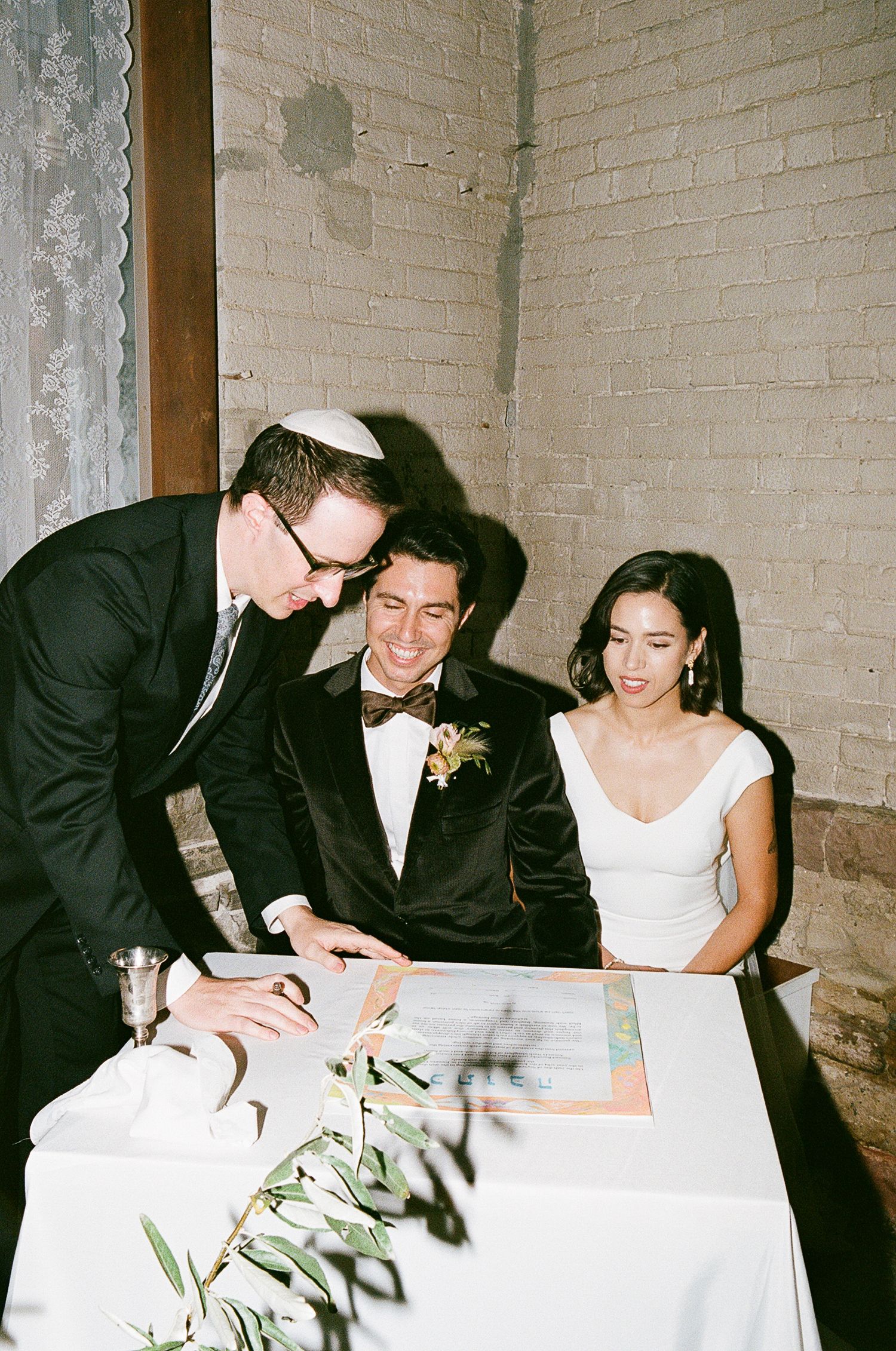 Three wedding guests sign documents at a white-clothed table during a formal ceremony.