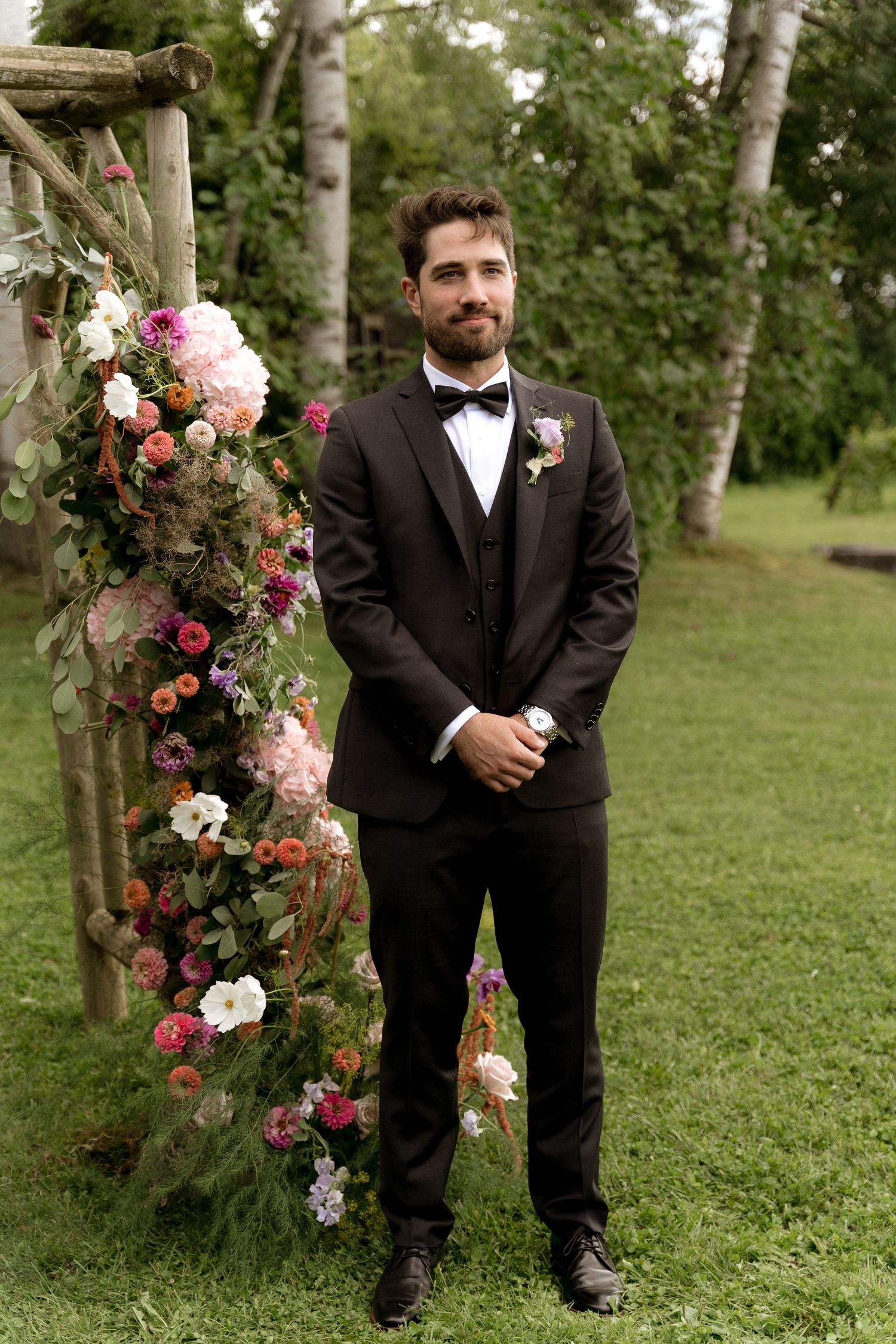 An elegant groom in a black tuxedo stands next to a wooden arch decorated with pink and white flowers in an outdoor garden setting.