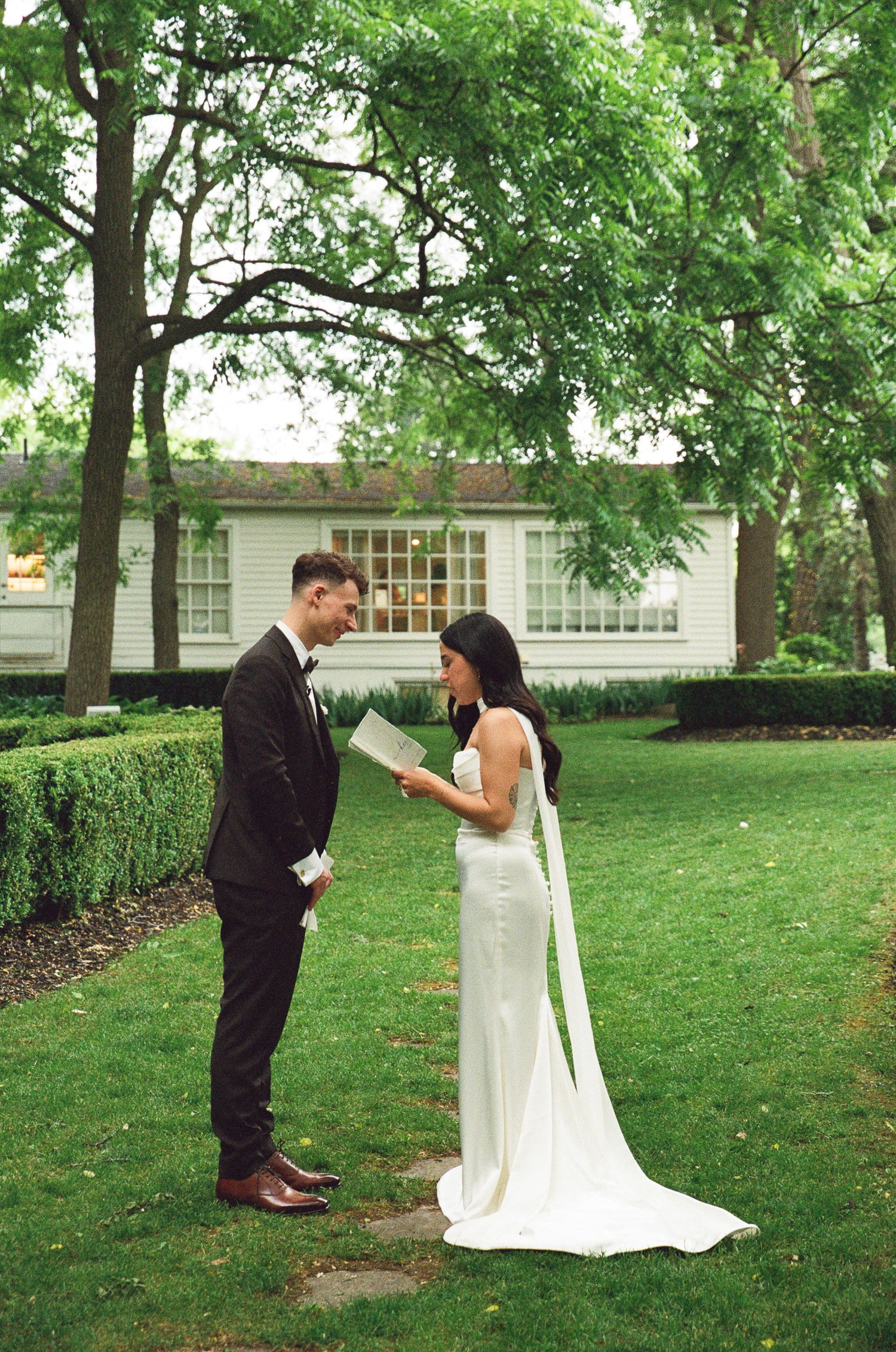 A couple shares a romantic moment on their wedding day in a lush garden setting with a white house in the background.