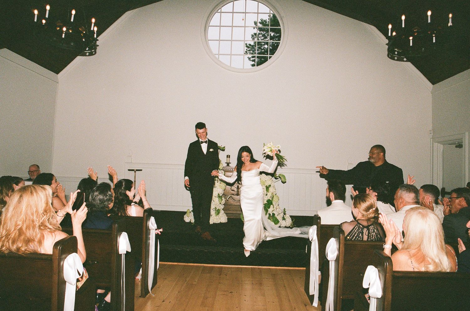 People gather in a small chapel with white walls and wooden pews for a wedding ceremony as candlelight glows overhead.