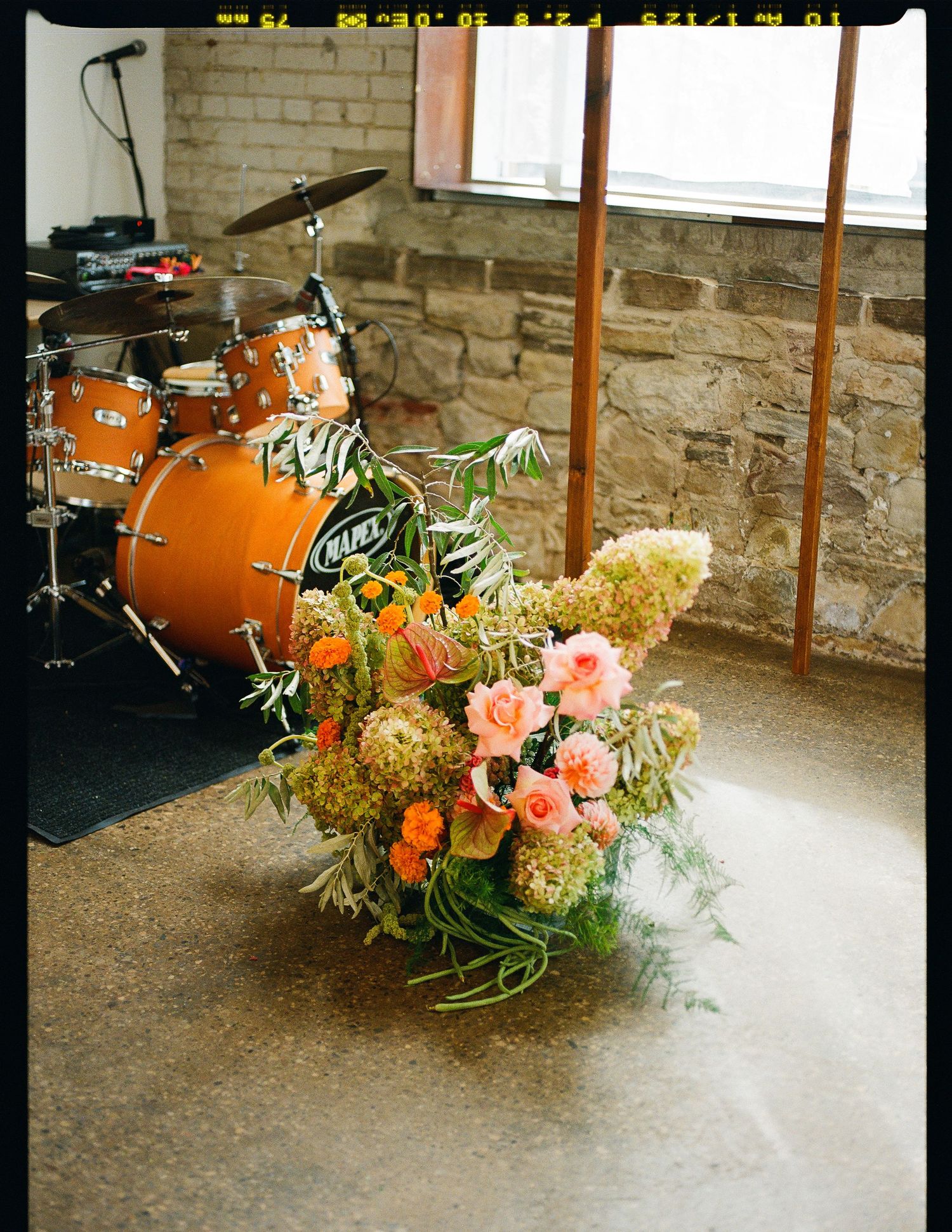 Orange drum kit and floral arrangement with peach roses and greenery against stone wall in natural lighting.