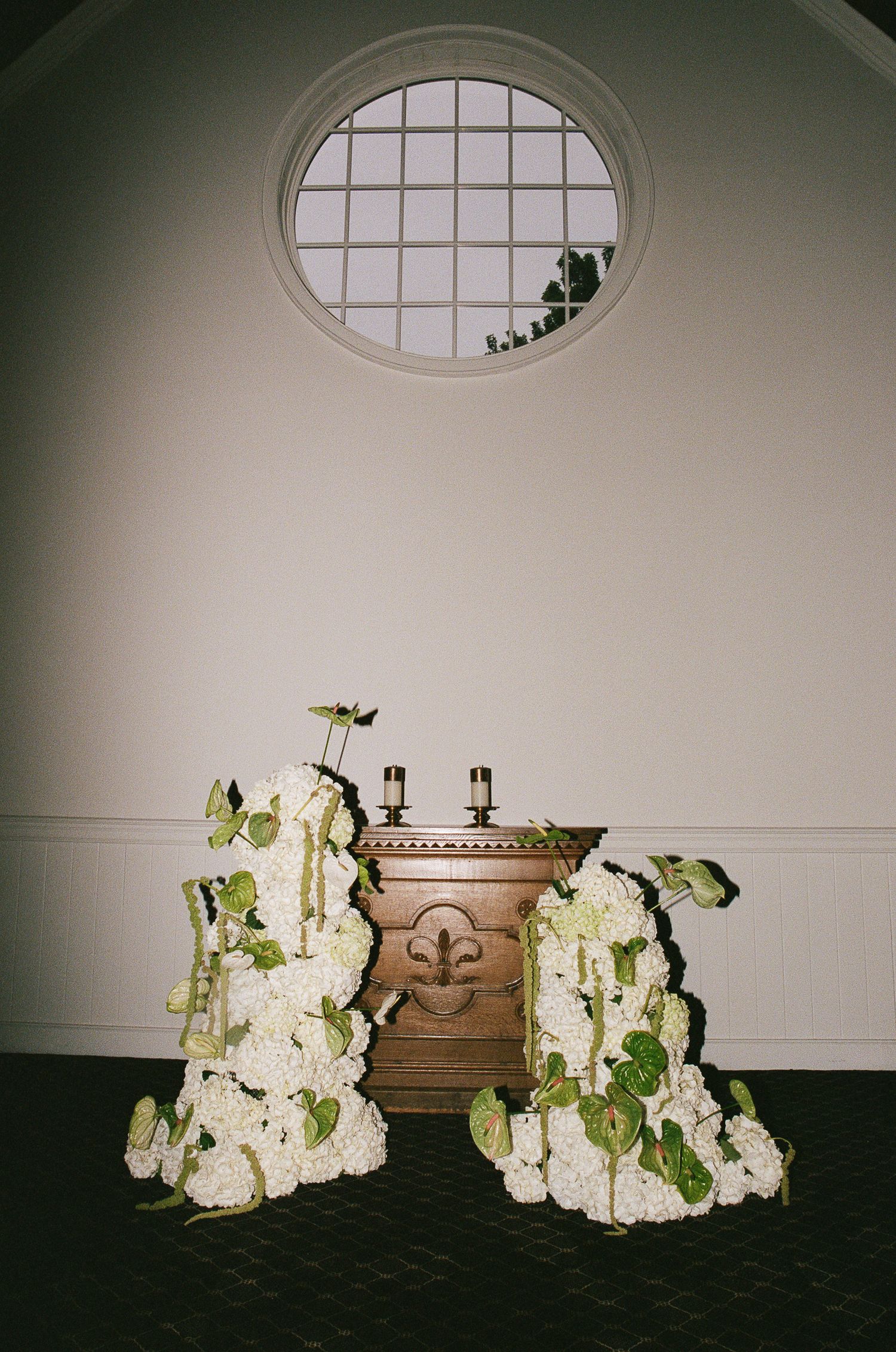 Elegant white floral arrangements sit below an oval window in dimly lit room with ornate wood furniture.