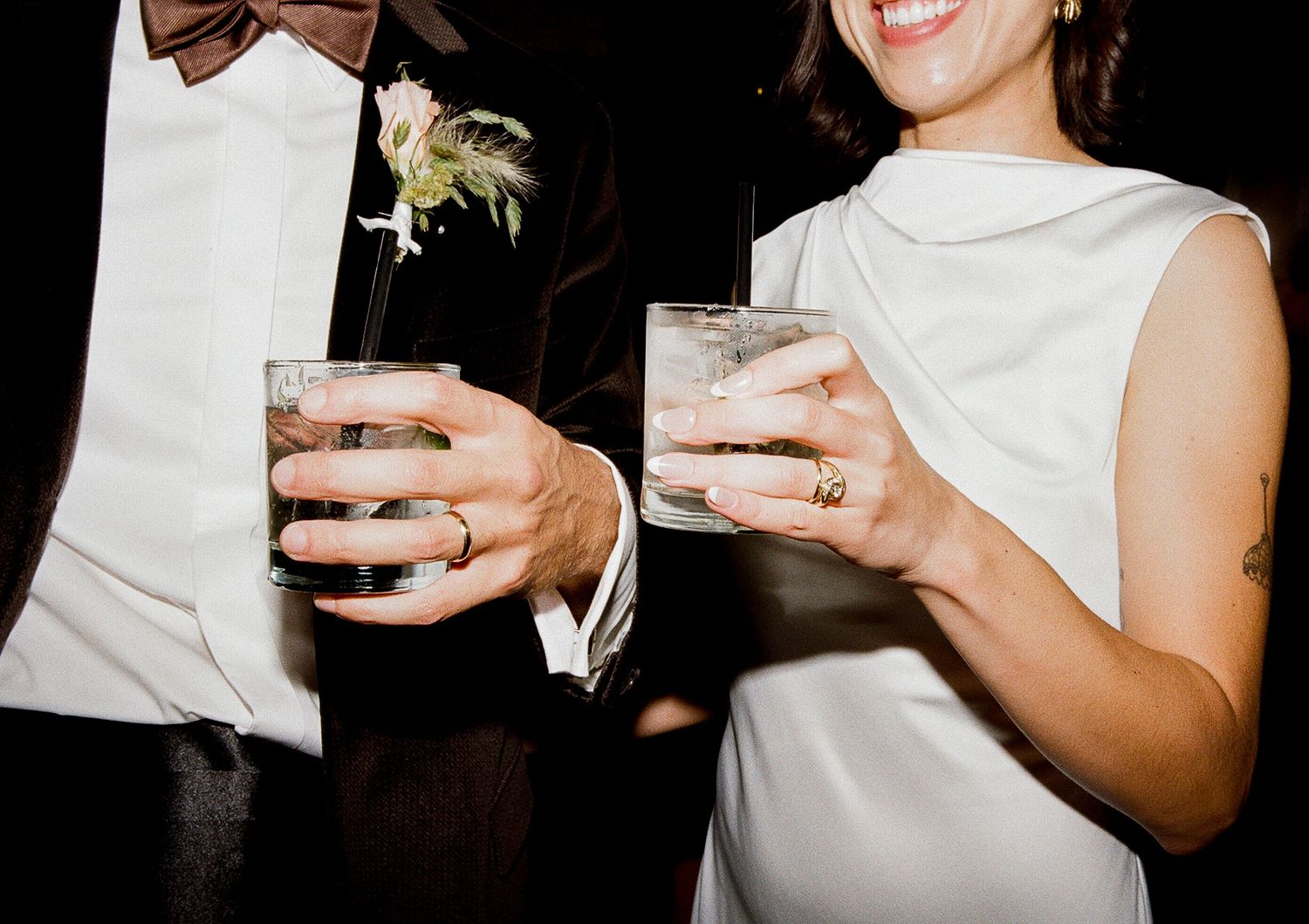 Close up of wedding guests holding drinks in formal attire, focusing on their hands and cocktail glasses.