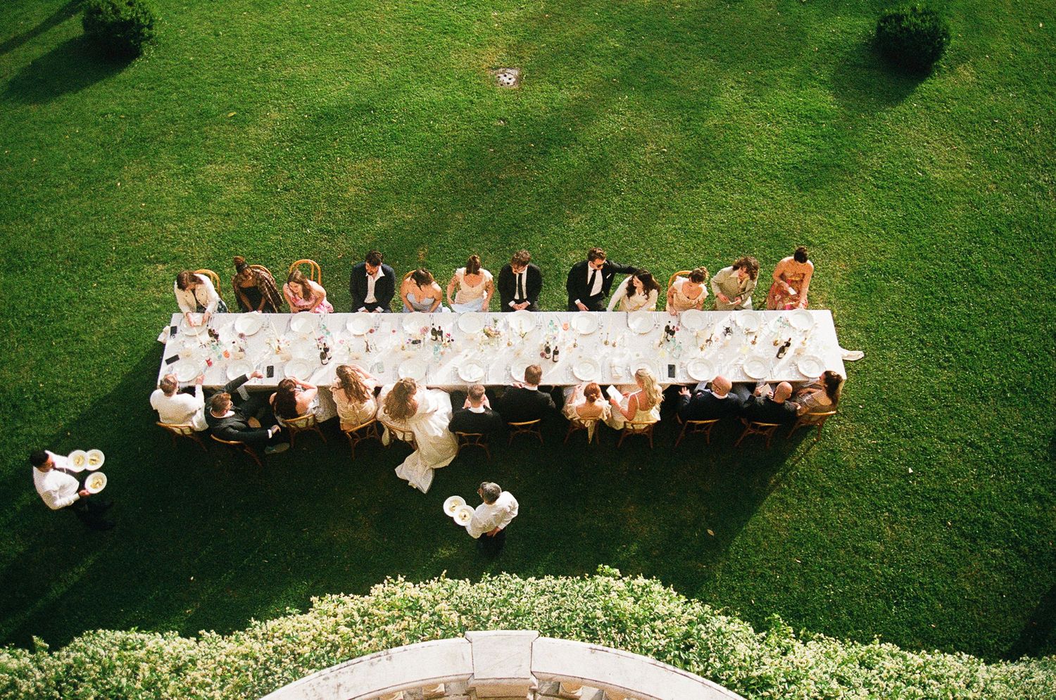 Aerial view of an outdoor wedding reception with a long white banquet table setup on a green lawn.