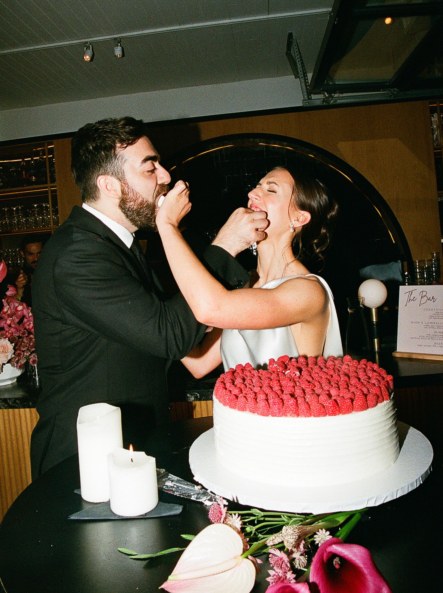 A couple playfully feeds each other wedding cake at their reception with a red-topped white cake and candles on the table.