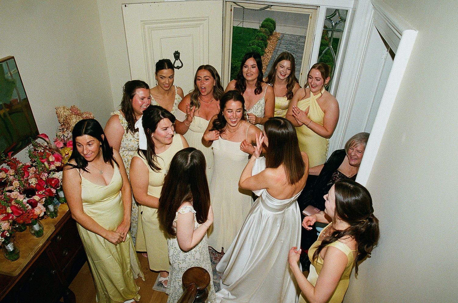 A group of wedding party guests gathered in light pastel dresses smile and celebrate in a cozy indoor setting.