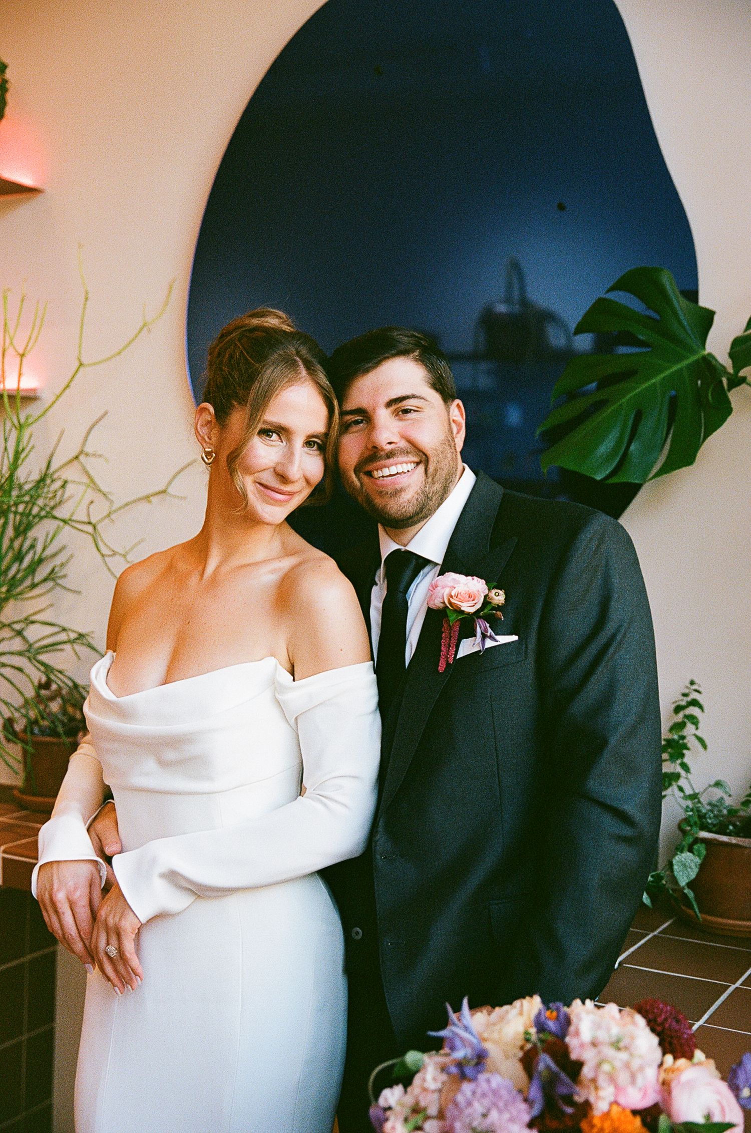 A couple poses together at their wedding, the bride in an elegant white off-shoulder gown and the groom in a dark suit.