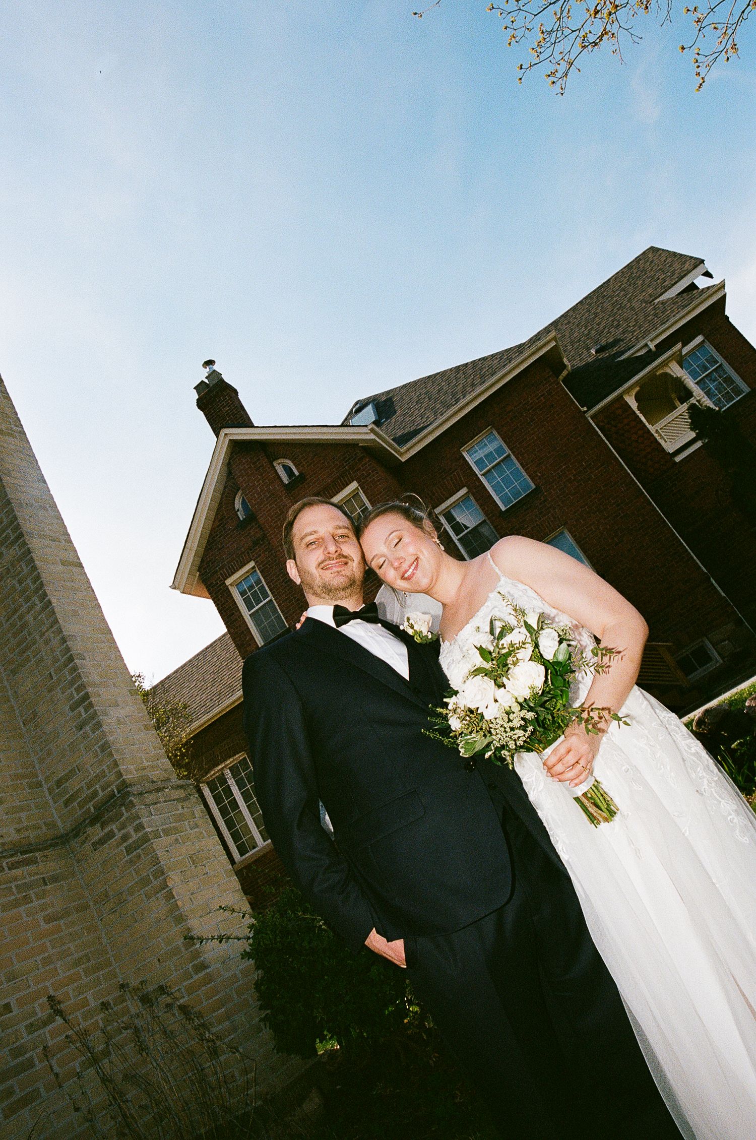 A joyful couple on their wedding day posing outside a brick house with a beautiful bouquet of white flowers.