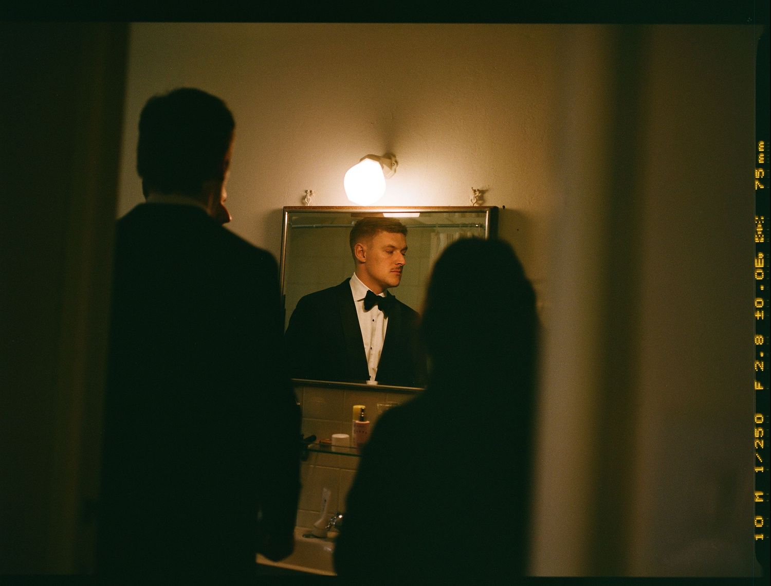 A speaker in a tuxedo stands at a podium in a dimly lit room while silhouetted figures listen in the foreground.