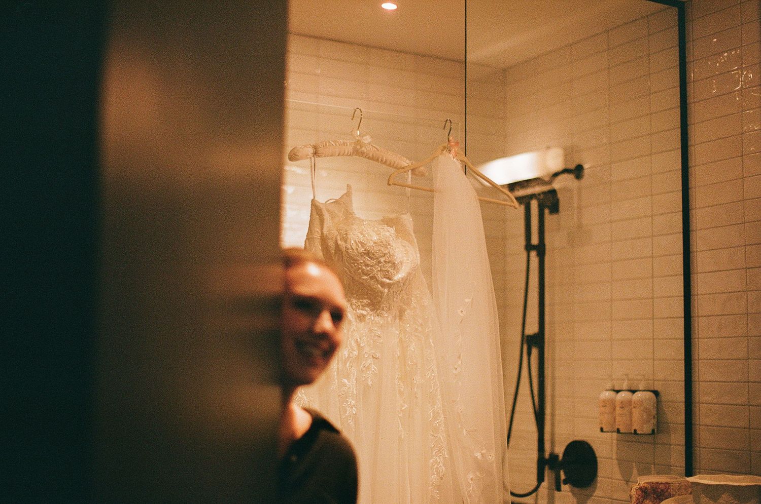 A bathroom interior with cream subway tiles showing a shower head and hanging white garments in warm lighting.