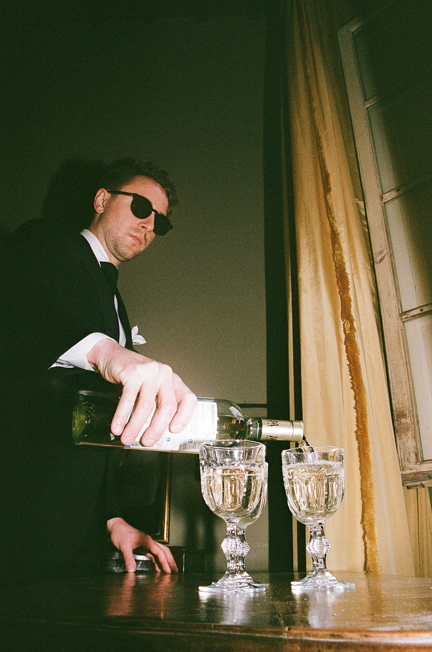 A person in sunglasses and formal attire pours champagne into crystal glasses near a window.