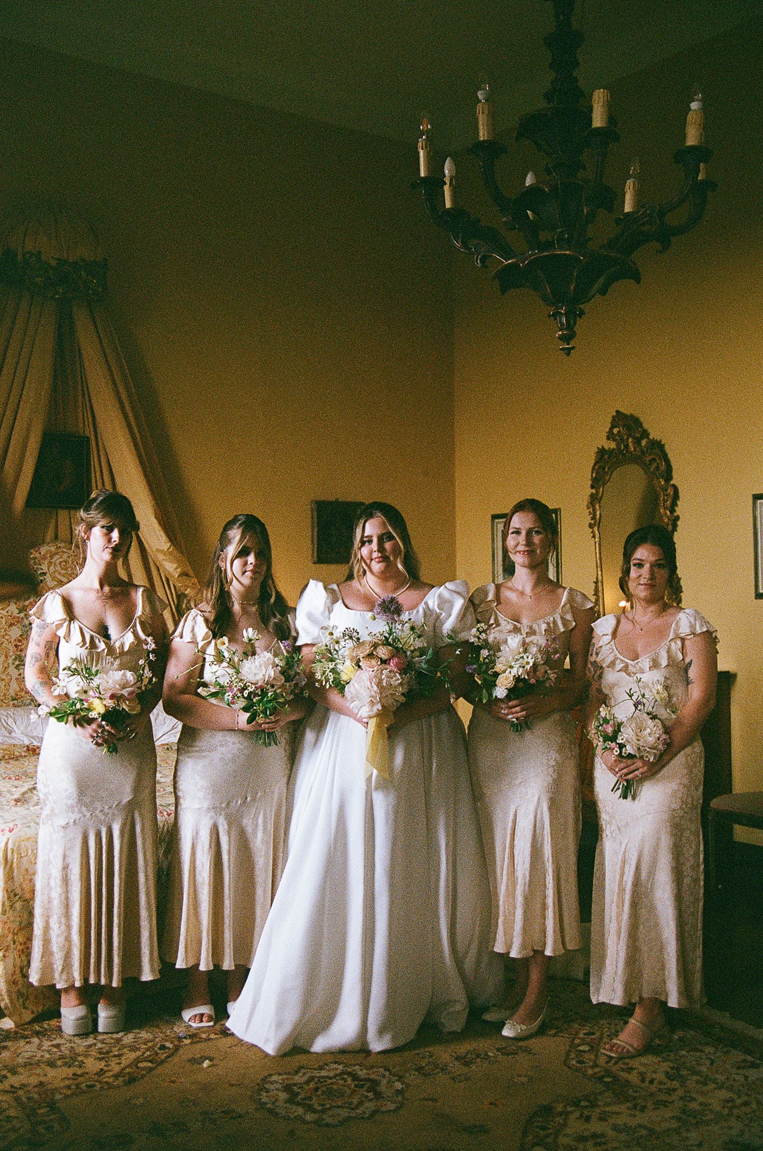 A bridal party wearing soft pastel dresses holds matching floral bouquets while standing in an elegant indoor setting.