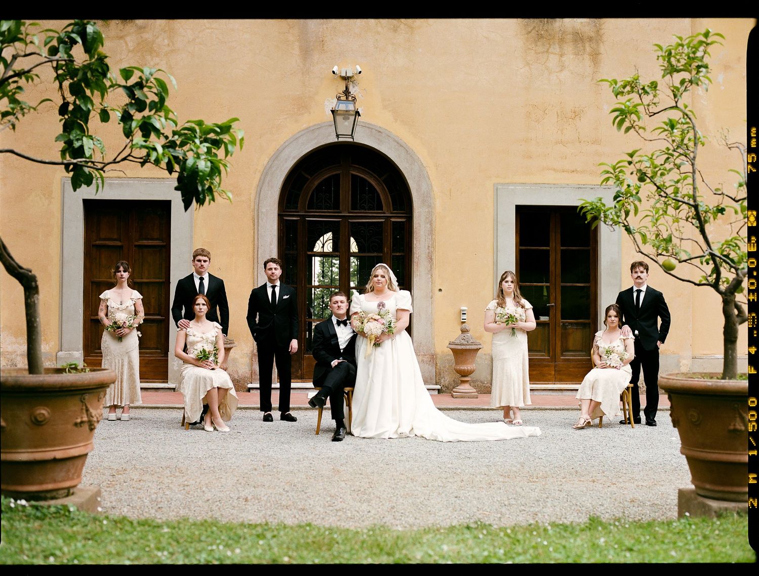 Wedding party poses elegantly on stone steps in front of arched doorway of classic building.