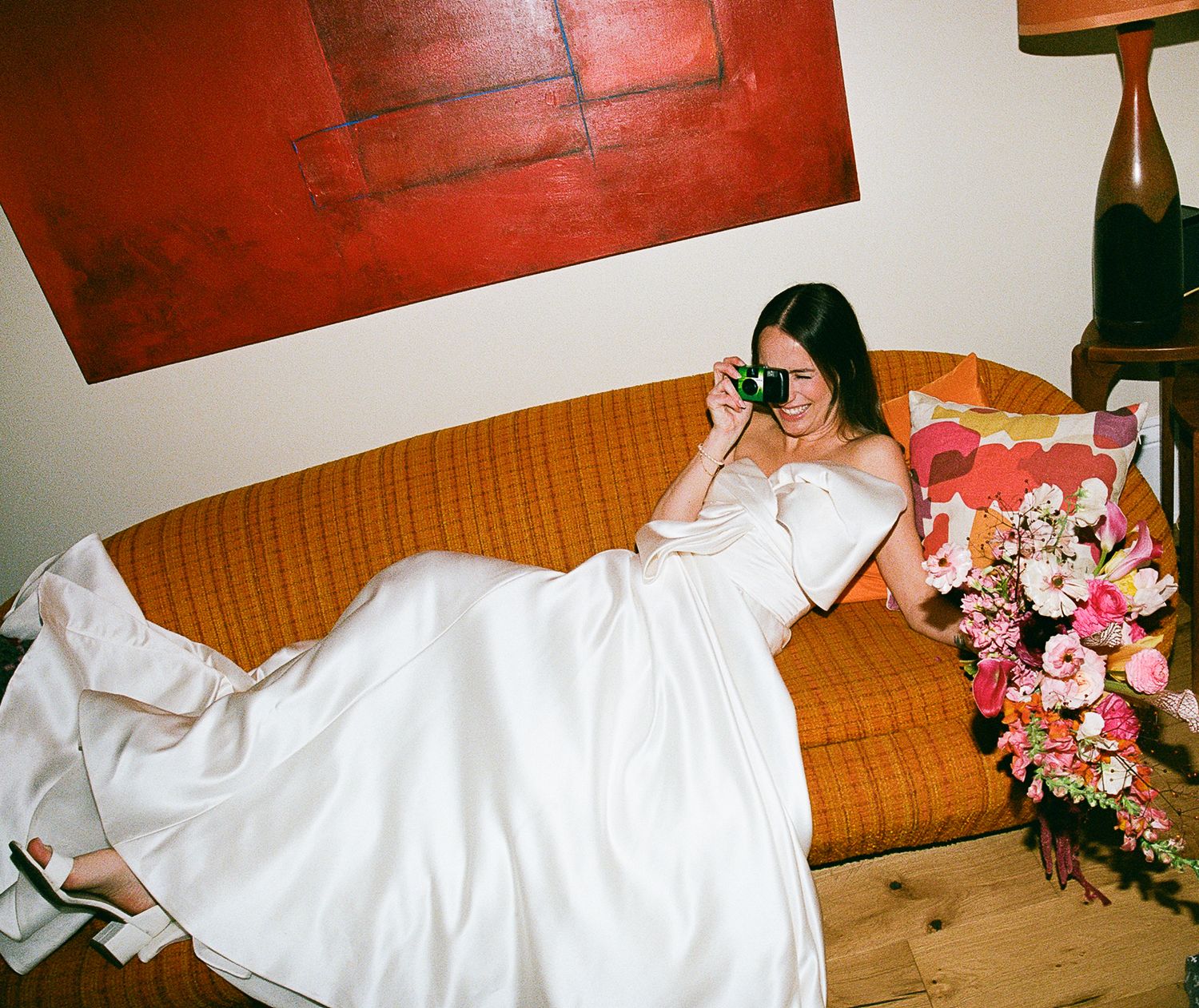A person in sunglasses reclines on a vintage orange couch wearing a flowing white dress with floral arrangements nearby.