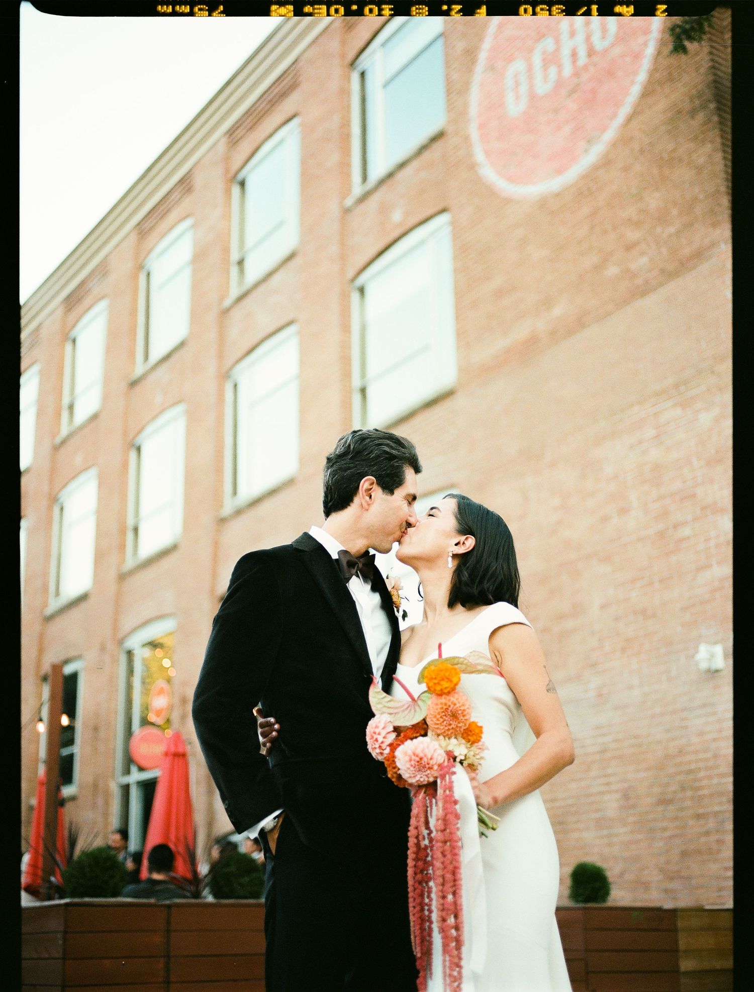 Wedding couple embrace in formal attire against brick industrial building with vintage film aesthetic.