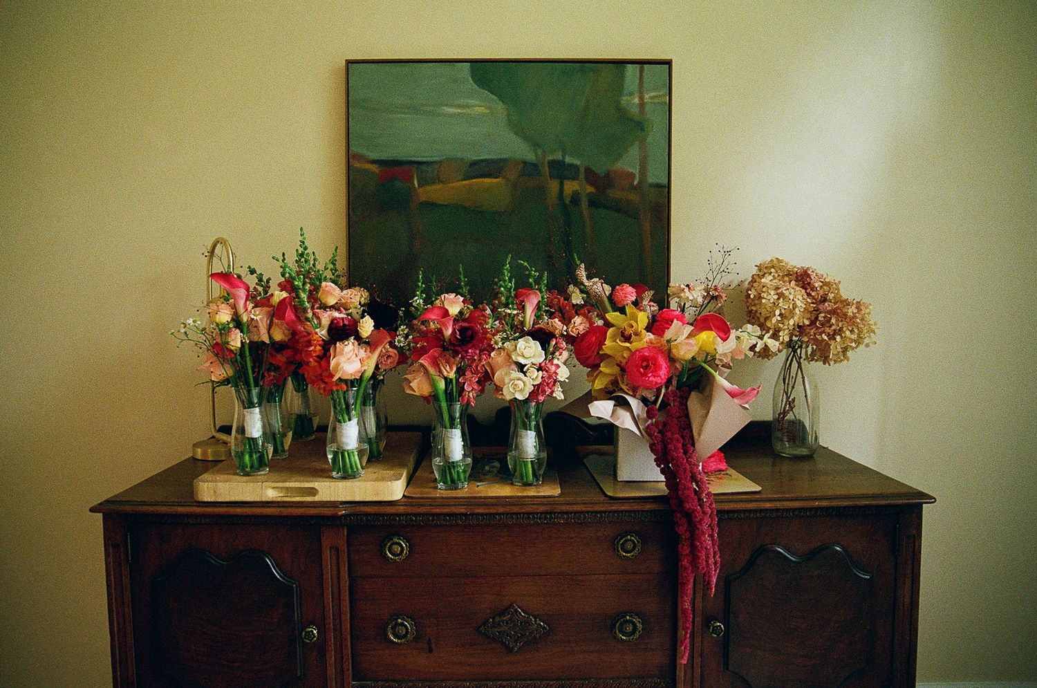 Multiple glass vases filled with vibrant red and pink flowers arranged on top of a dark wooden dresser against a green wall.
