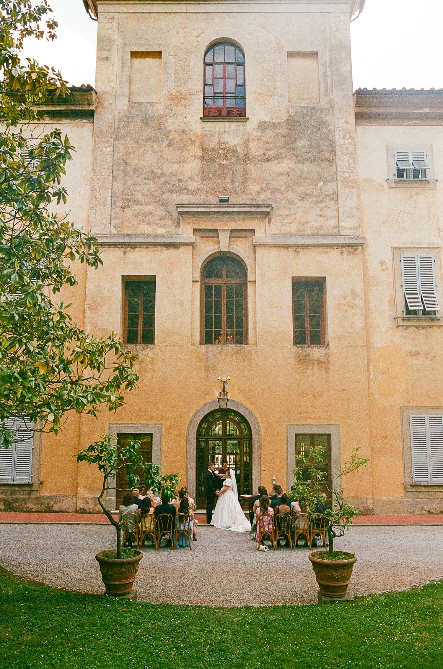 Historic villa with arched entrance hosts outdoor wedding ceremony with potted plants framing the entrance.