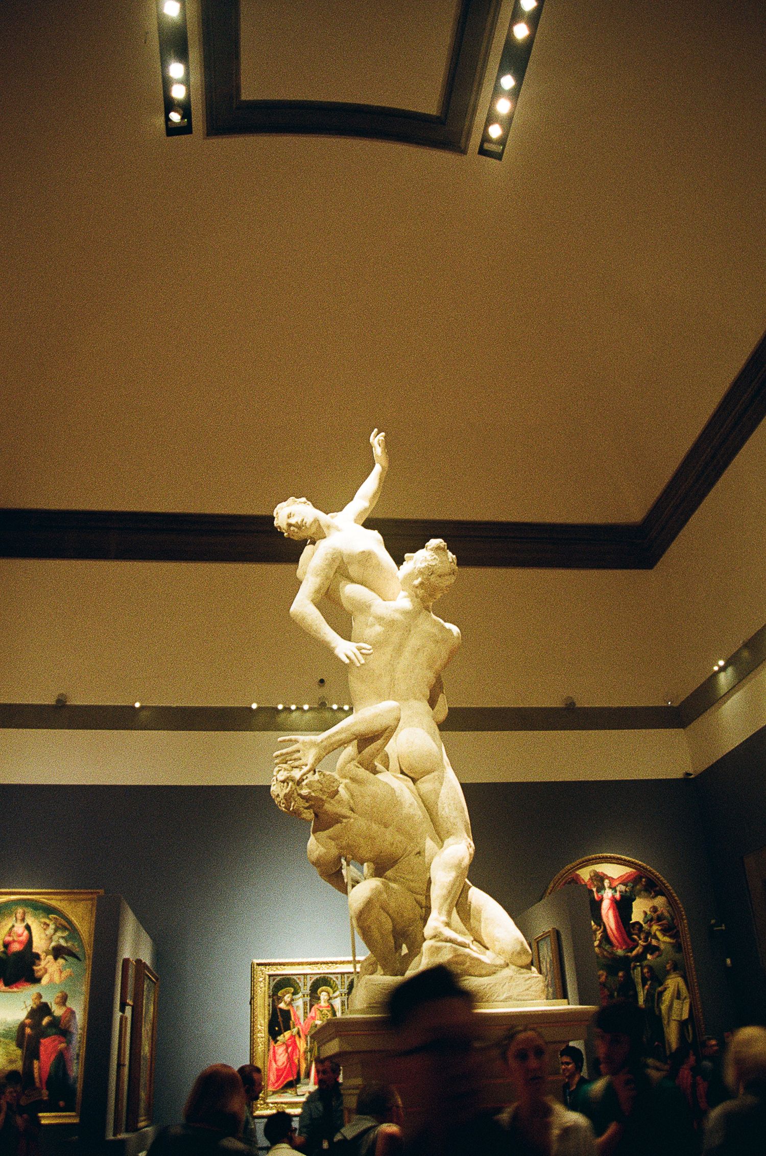 A dramatic marble sculpture displayed in a museum gallery shows two entwined figures in an upward-reaching pose.