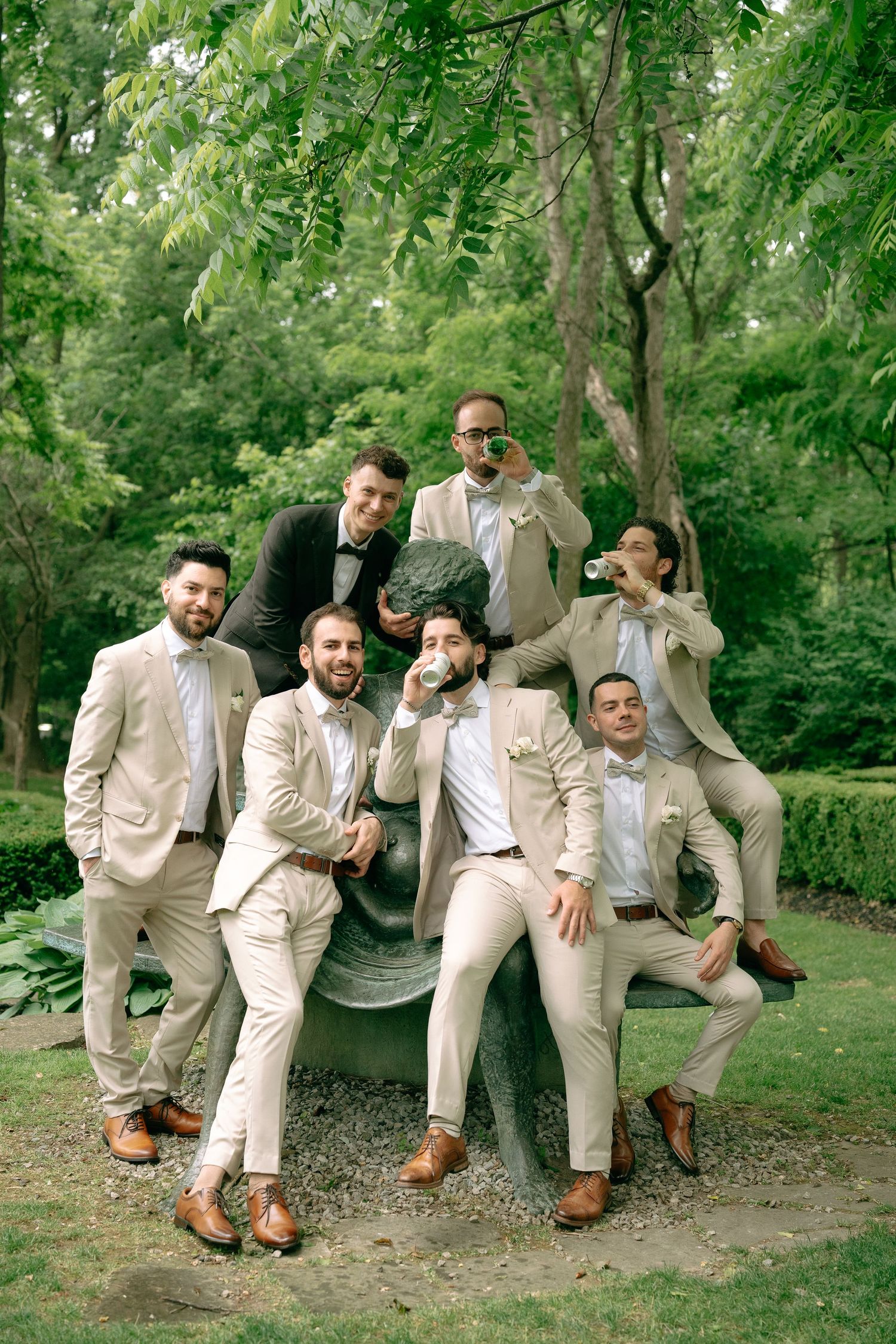Wedding party wearing matching beige suits pose playfully together in a lush garden setting.