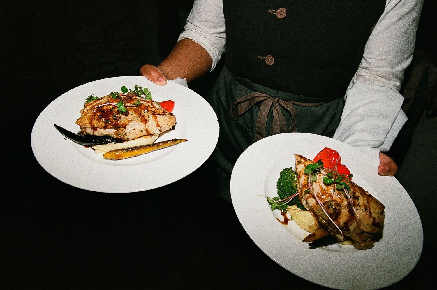 Two elegantly plated dishes of grilled meat and vegetables being served by a waiter.