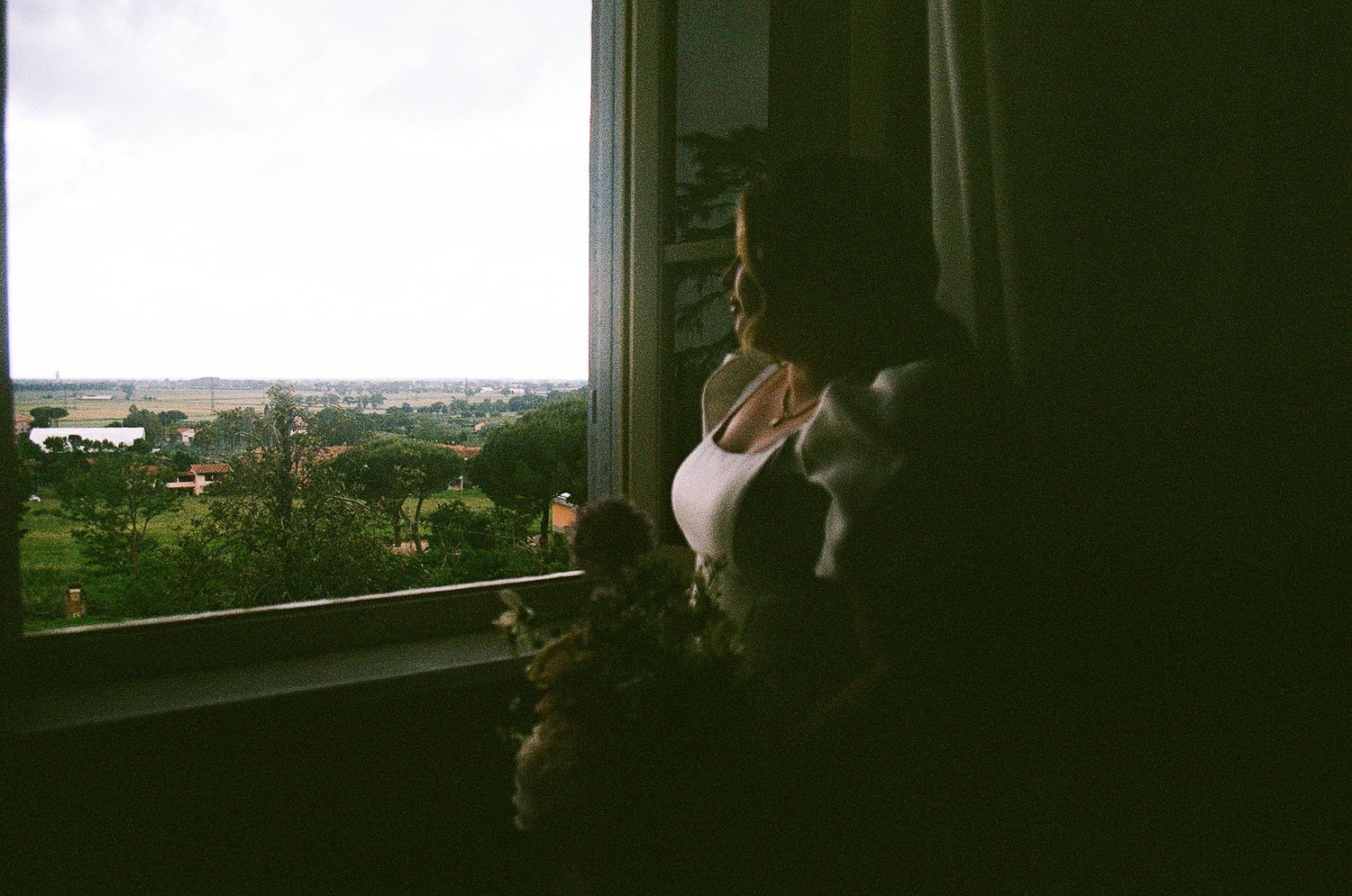 Looking out through a window at a distant cityscape, casting a moody silhouette against the natural light.