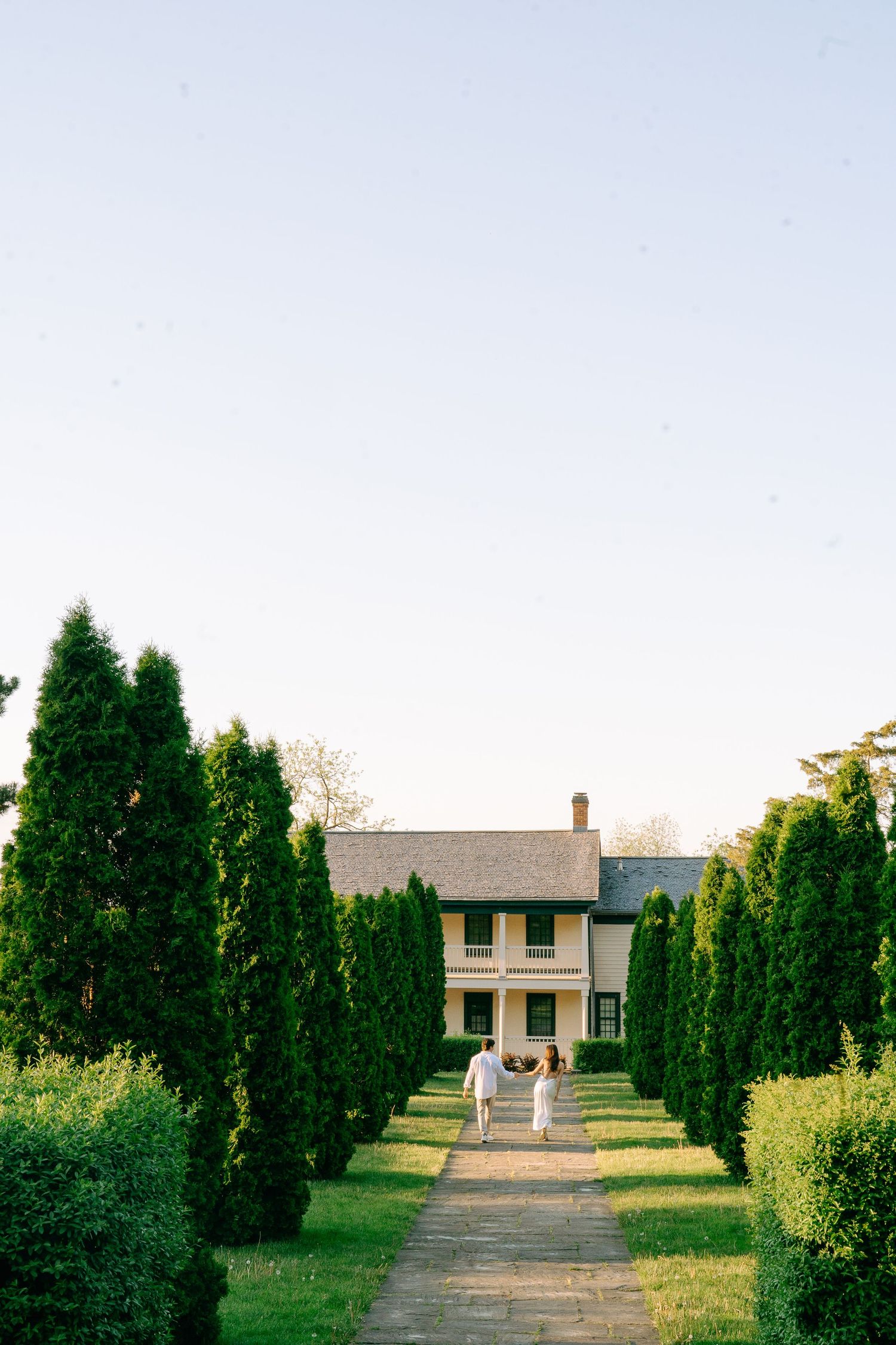 A white farmhouse with green trees lining a stone pathway leading to the entrance at sunset.
