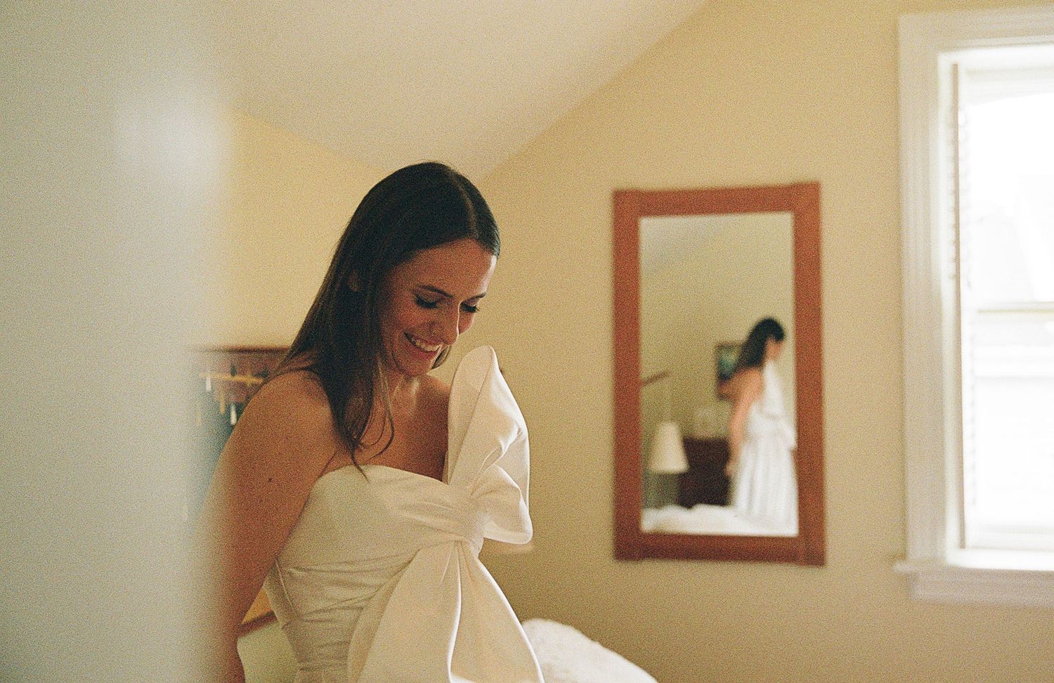 Woman in white wedding dress looks down while getting ready in sunlit dressing room with mirror.
