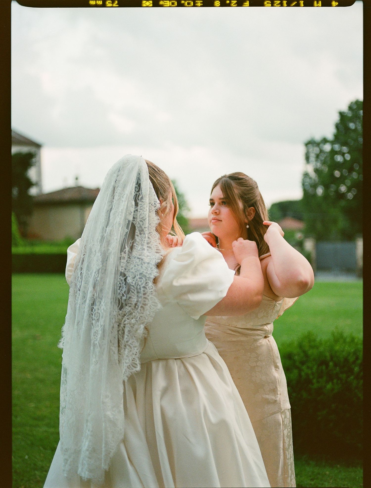A bride in a white gown and veil stands with another person in beige on a grassy lawn during overcast weather.