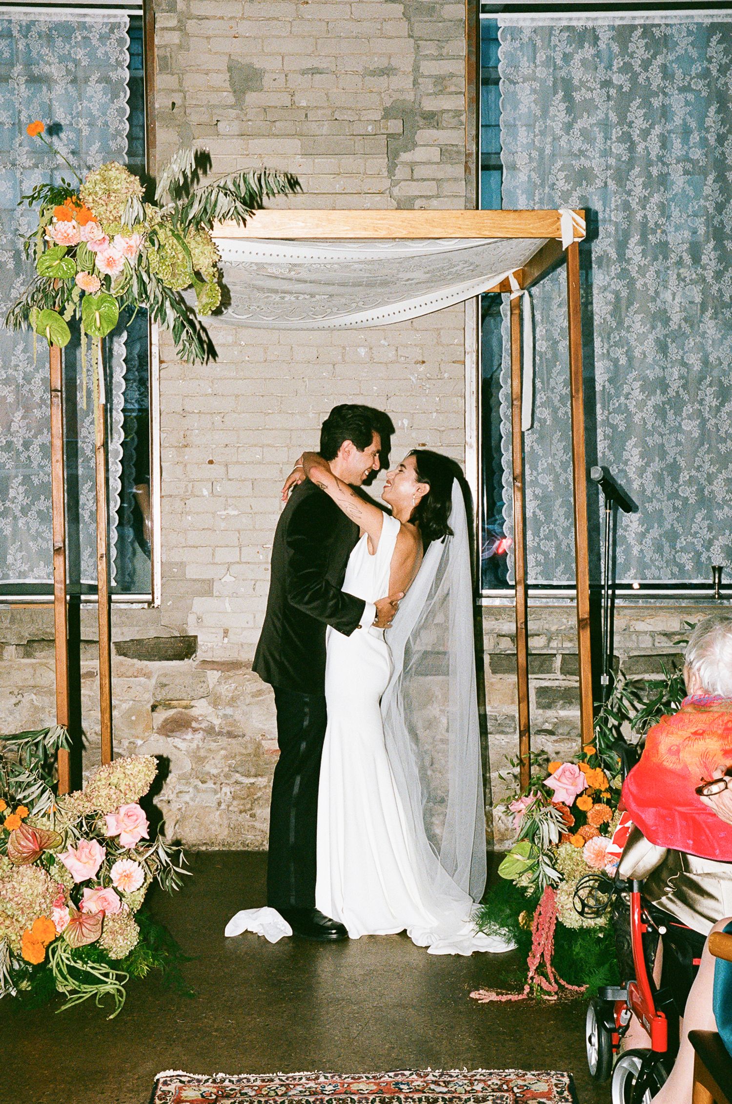 Wedding ceremony under a floral-decorated canopy arch with white draping against exposed brick wall.